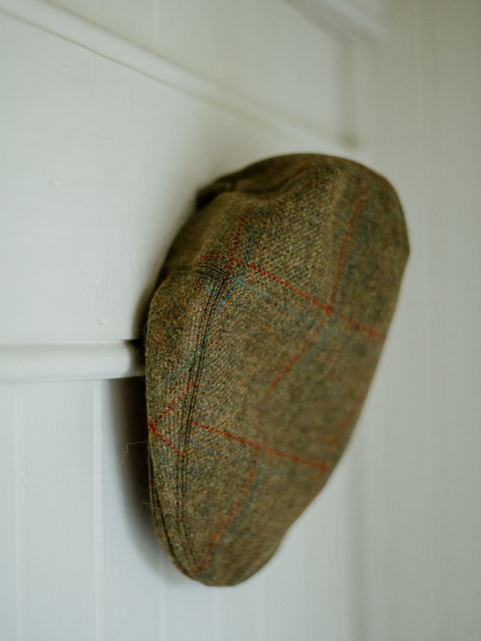 The Mini Tweed Cap by Campbell's of Beauly, with its brown checkered design, hangs on a white wooden wall—an ideal way to add country charm to your child’s outfit.