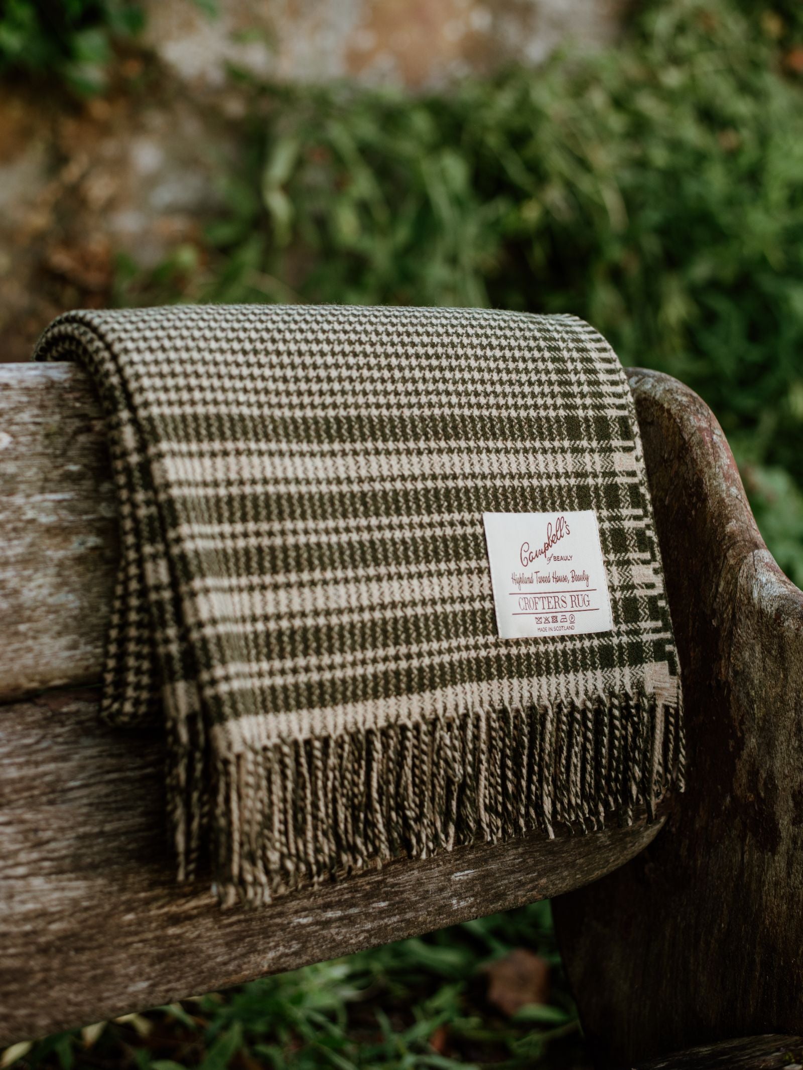 A Crofters Blanket by Campbell's of Beauly with fringed edges is draped over a wooden bench outdoors. Green foliage and a blurred background enhance the cozy country aesthetic of the scene.