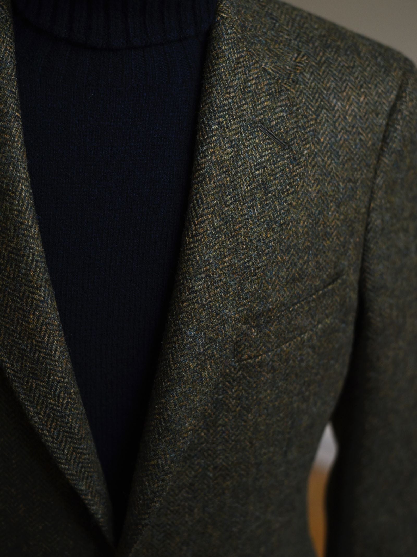 Close-up of a person wearing the Campbell's of Beauly Auld Stock Green Herringbone House Jacket over a black ribbed turtleneck. Only the upper body is visible, highlighting the textured tweed fabric and herringbone pattern.