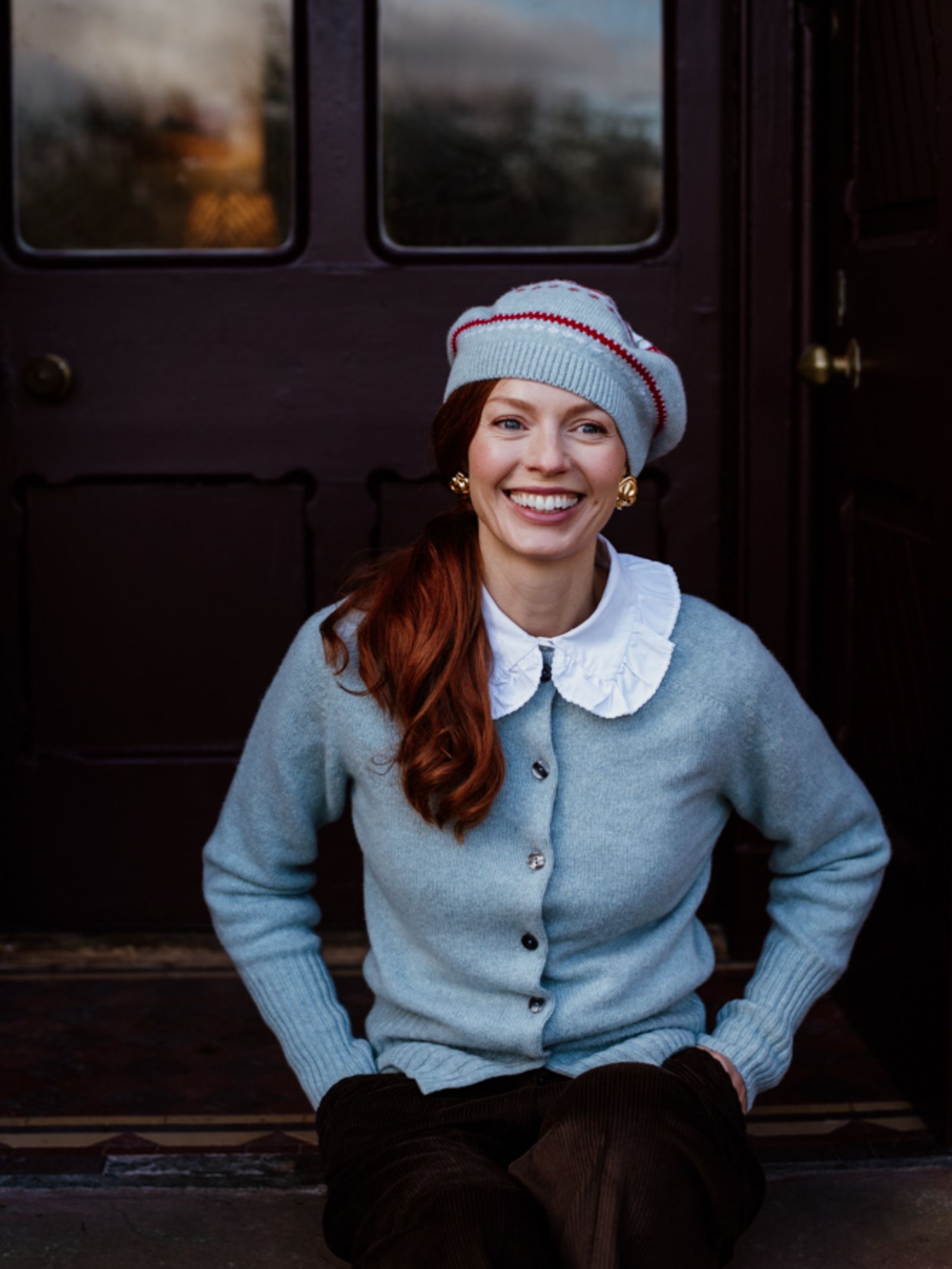 A smiling woman with long red hair, wearing a light blue cardigan, white ruffled shirt, and the Campbells of Beauly Strawberry Beret, sits on a doorstep in front of a dark wooden door.