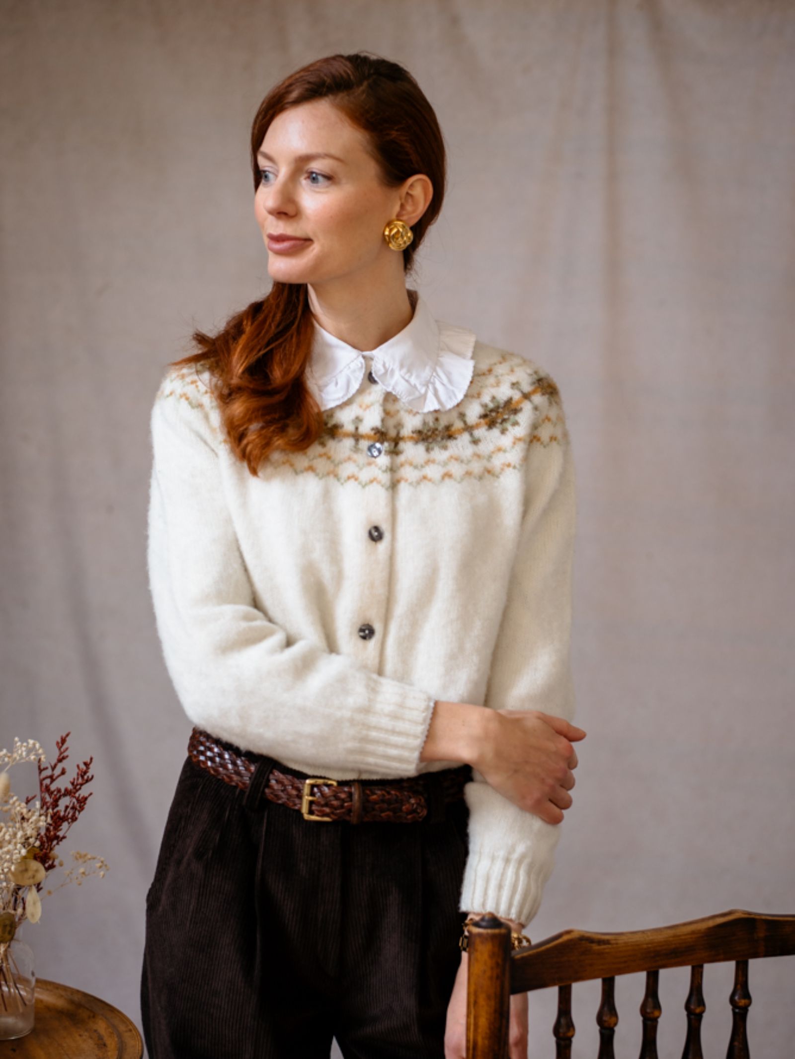 A woman with red hair wears the Campbell's of Beauly Brushed Shetland Fairisle Cardigan, featuring a ruffled white collar, dark brown corduroy pants, and a braided brown belt.