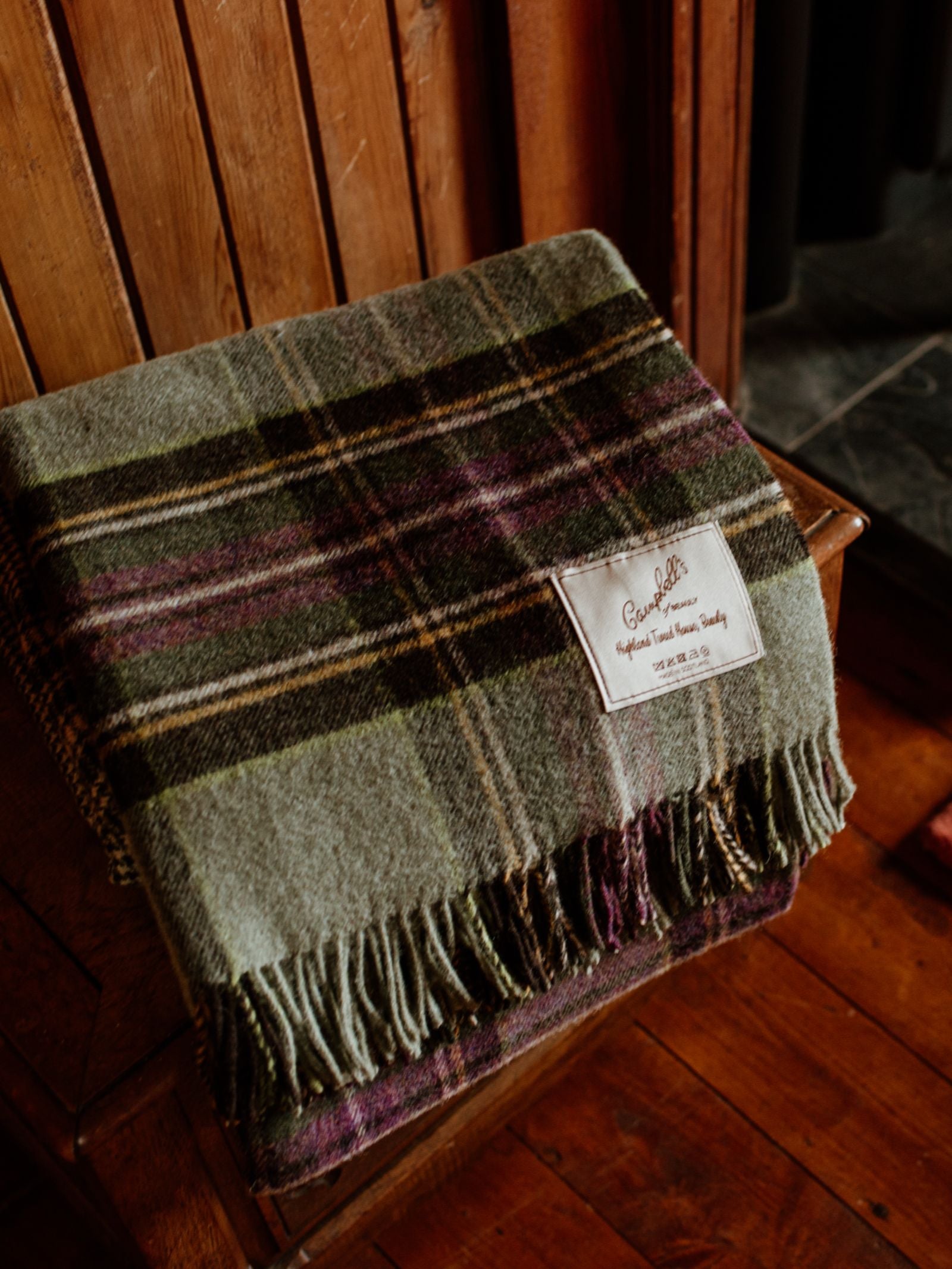 The Campbell's of Beauly Strathglass Throw, a green and purple plaid lambswool blanket with fringe and white label, is neatly folded on a wooden bench—a warmly lit scene for cosy home accessory lovers.