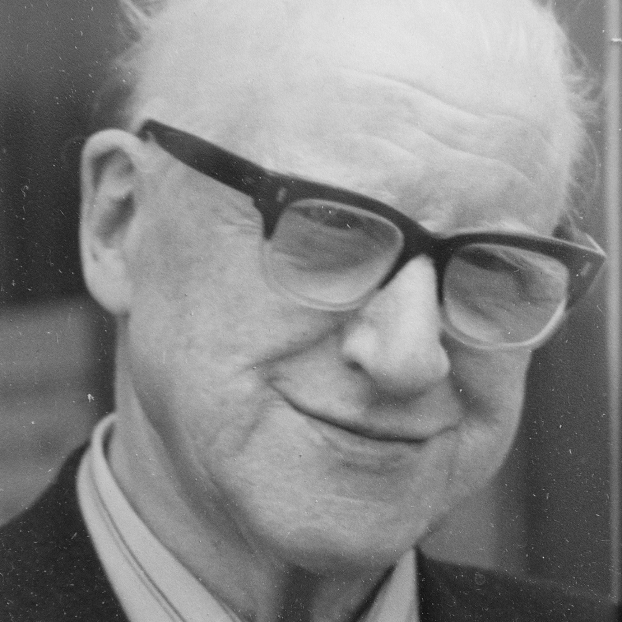 Elderly man with light skin, white hair, and wearing dark-rimmed glasses. He is smiling gently and dressed in a collared shirt and suit jacket. The photo is black and white and closely cropped to his face.