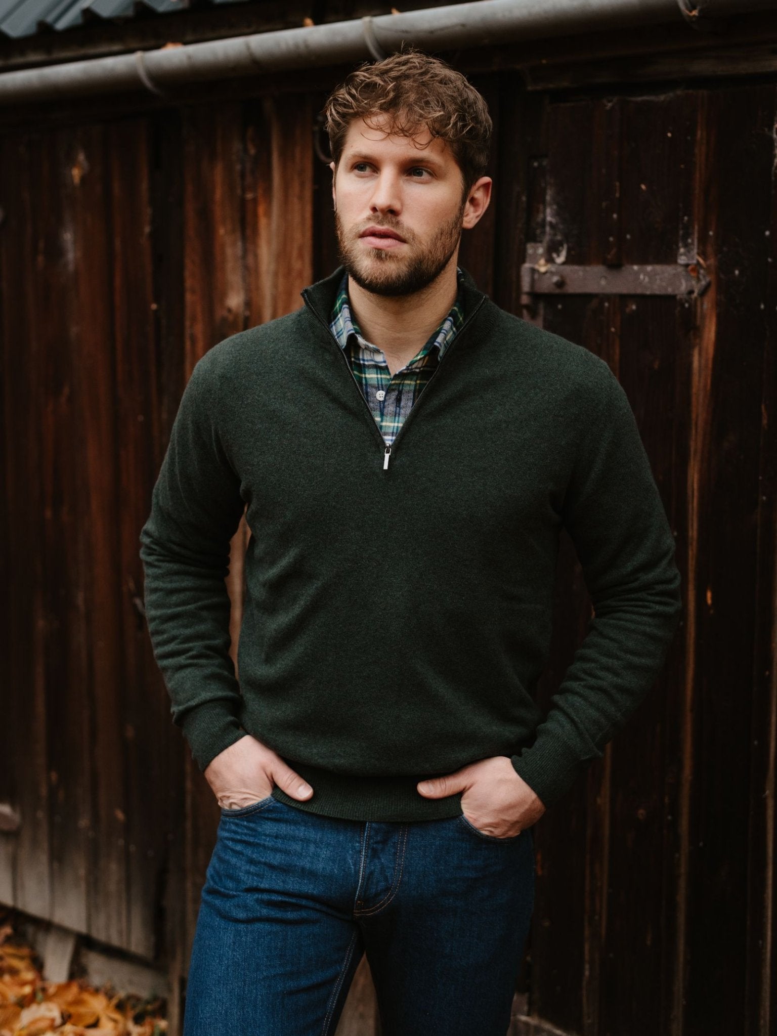 A man with short brown hair and a beard stands before a rustic wooden wall, wearing Campbell’s of Beauly’s Merino & Cashmere Quarter Zip Jumper in dark green over a plaid shirt and blue jeans, hands in pockets, looking slightly to the side.