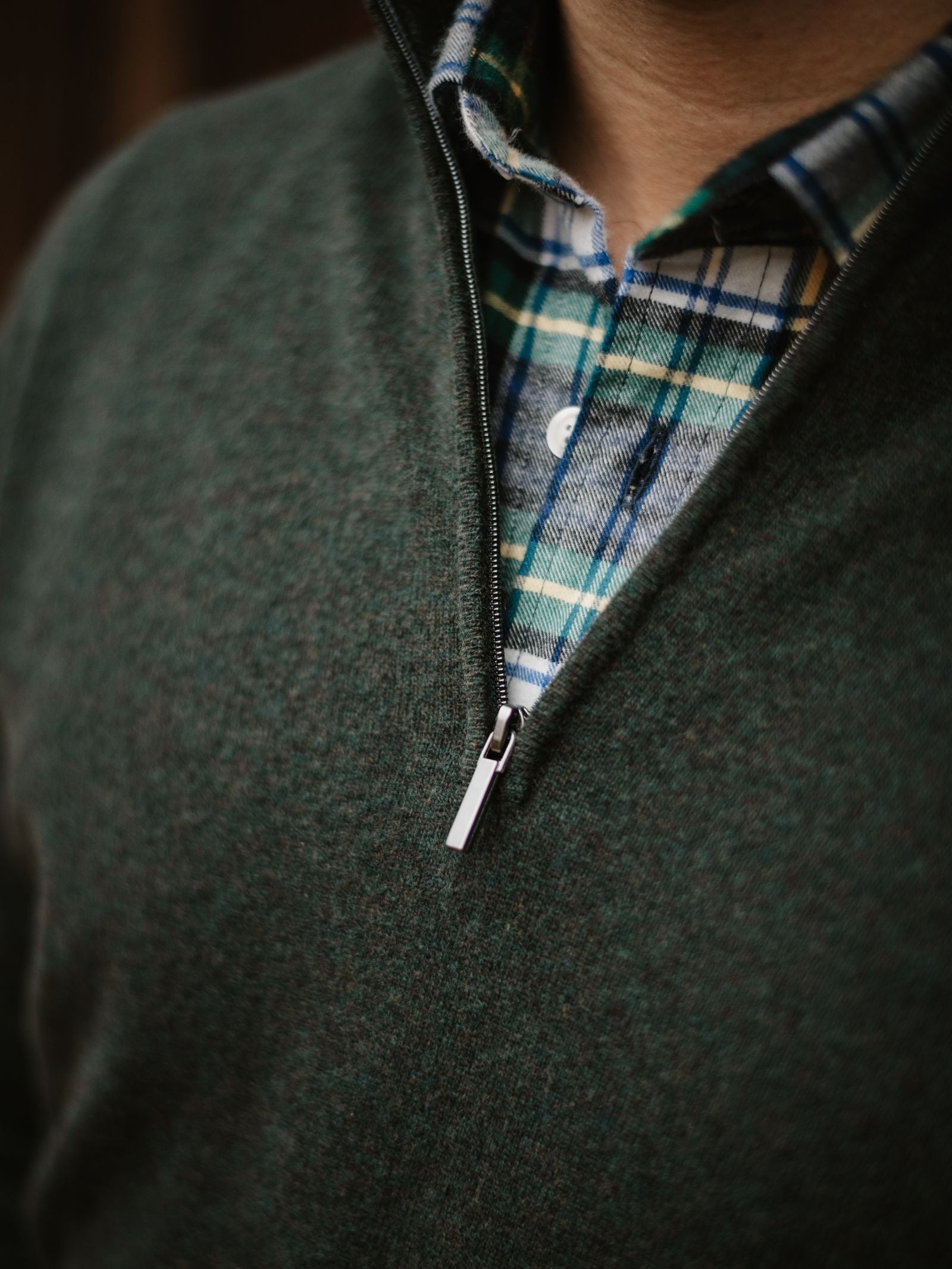 Close-up of a person wearing a green Campbell's of Beauly Merino & Cashmere Quarter Zip Jumper over a blue, green, and white plaid button-down shirt, with the zipper partially open to reveal the shirt underneath.