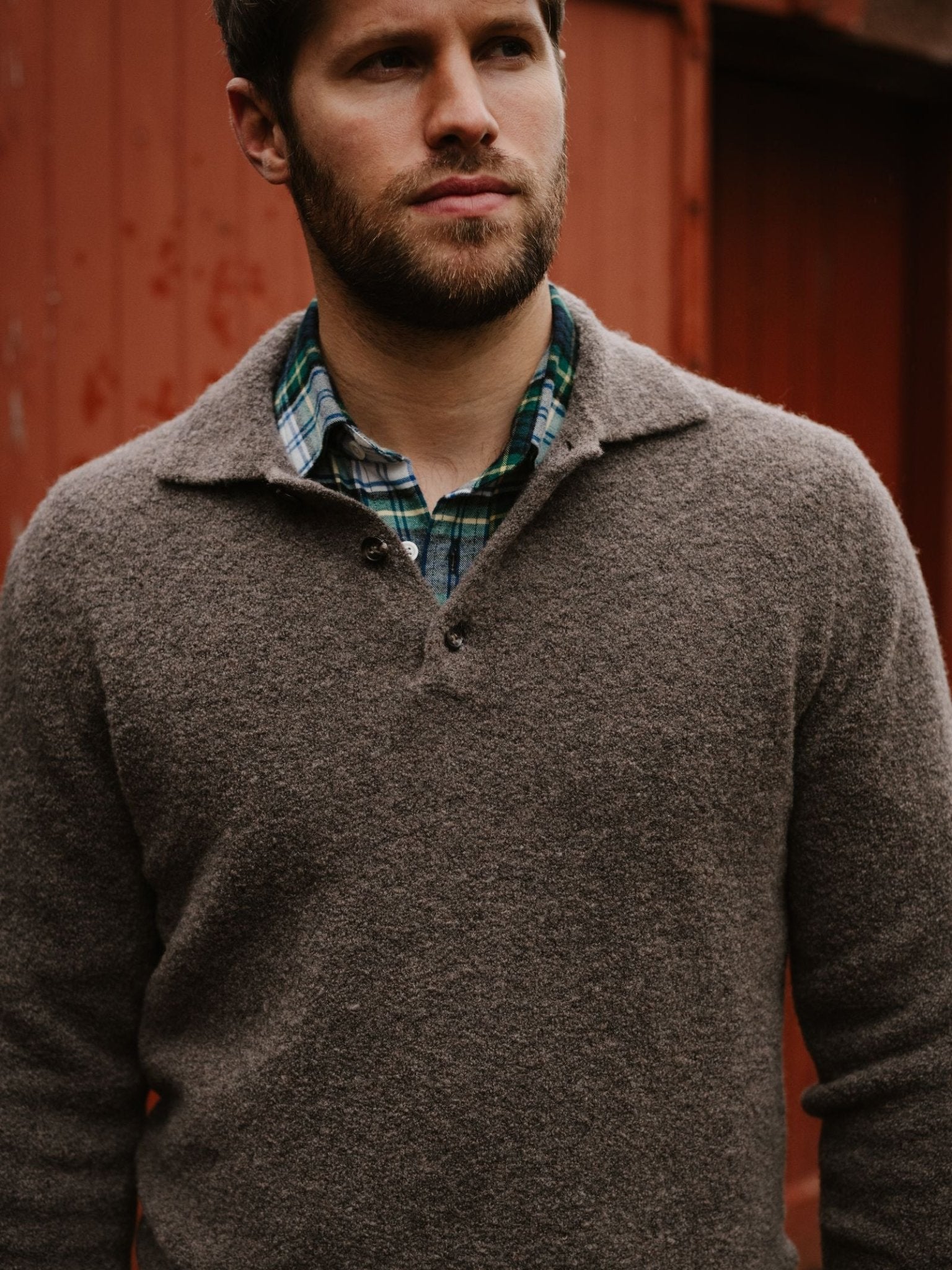 A man with a beard wears the Auld Stock Bouclé Neck Jumper by Campbell's of Beauly over a green and blue plaid shirt, standing in front of a red wooden wall.