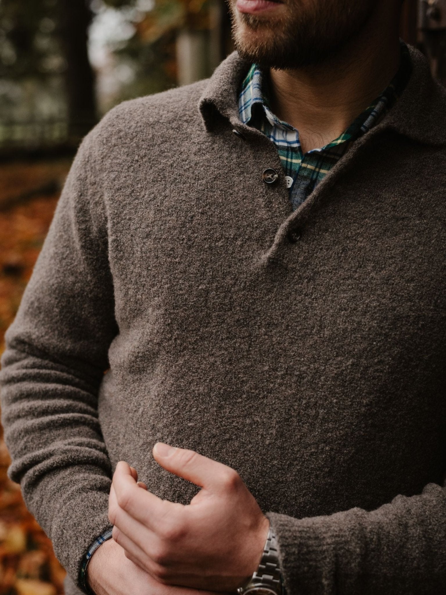 A man in the Auld Stock Bouclé Neck Jumper by Campbell's of Beauly, layered over a green and blue plaid shirt, stands outdoors in autumn with blurred fallen leaves in the background, visible from the chest up.