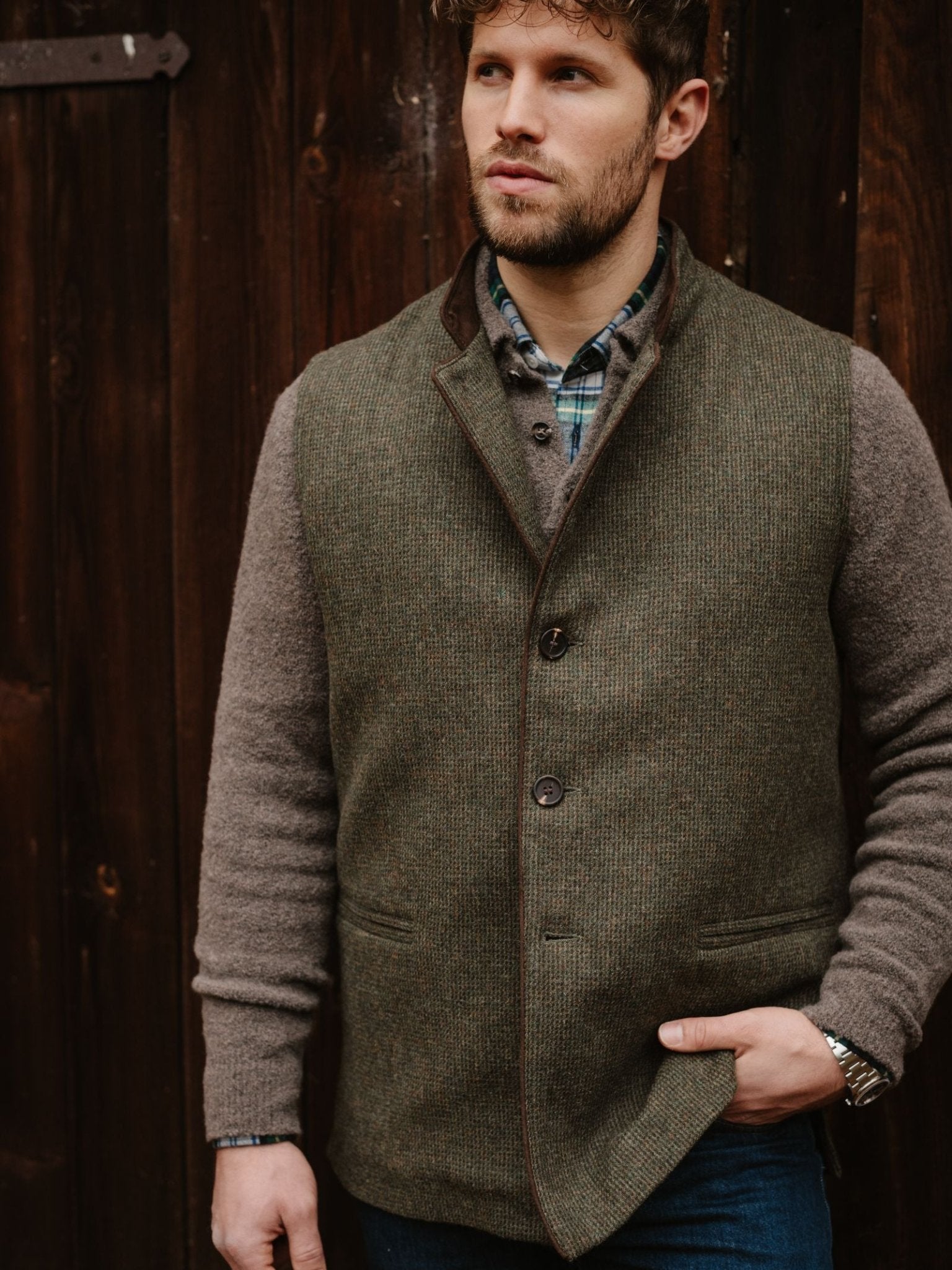 A man with brown hair and a beard stands before a wooden wall, wearing the Campbell's of Beauly Tweed Nehru Gilet over a brown sweater and green plaid shirt, one hand in his pocket, gazing to the side.