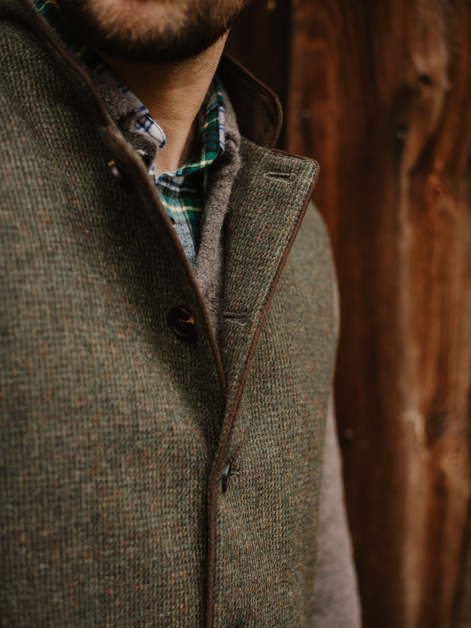 A man wears the Campbell's of Beauly Tweed Nehru Gilet over a gray sweater and green plaid shirt, standing before a wooden background. Only the lower half of his face and torso are visible.
