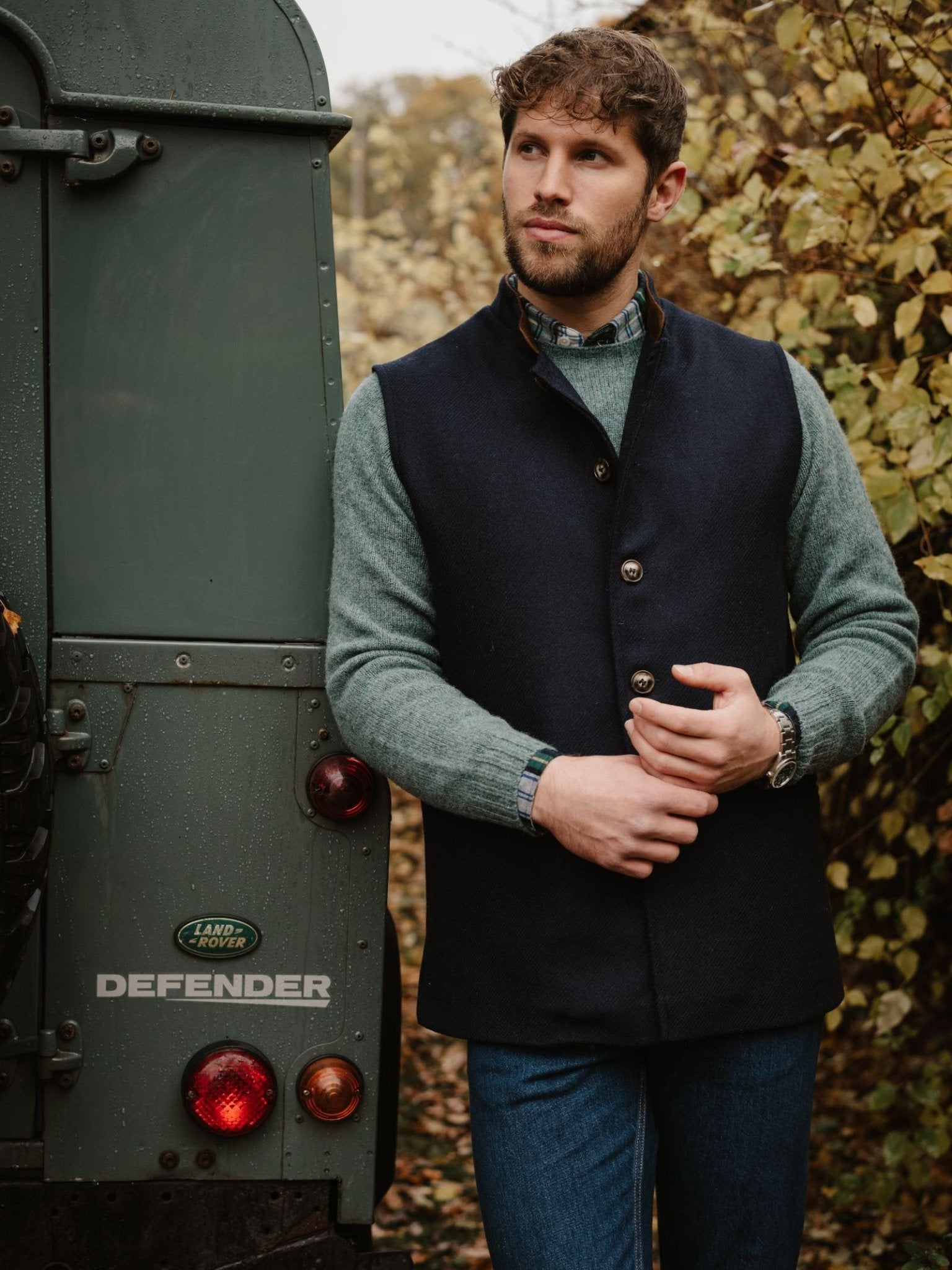 A man wearing a Campbell's of Beauly Tweed Nehru Gilet and green shirt stands by a green Land Rover Defender amid autumn foliage, gazing thoughtfully into the distance with one hand resting on his jacket.