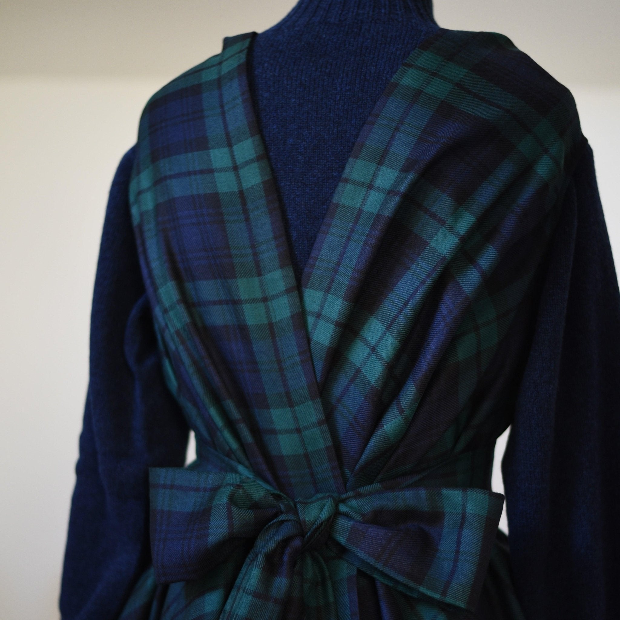 A close-up of a person wearing a dark blue sweater and a Campbell's of Beauly Reversible Wool Wrap Dress in green and navy tartan wool, tied with a large bow at the back against a plain light background.