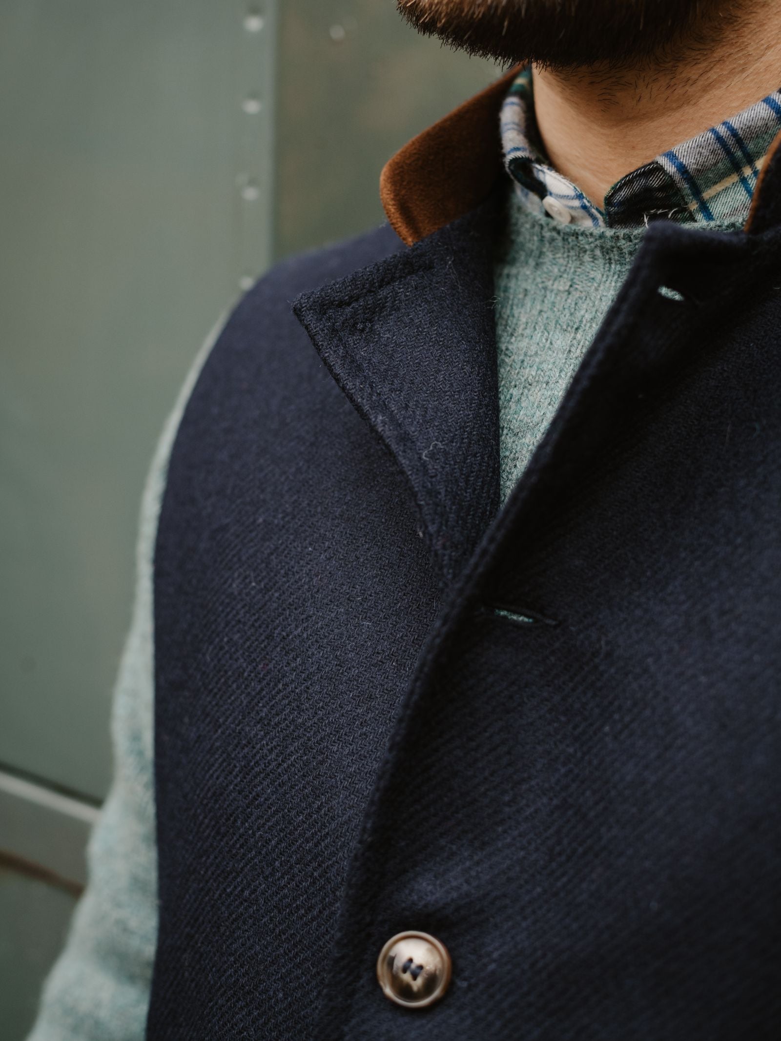 A close-up of a man wearing Campbell's of Beauly’s Tweed Nehru Gilet over a light blue sweater and plaid shirt, with a touch of brown collar and button; classic British style made in the UK. The background is blurred.