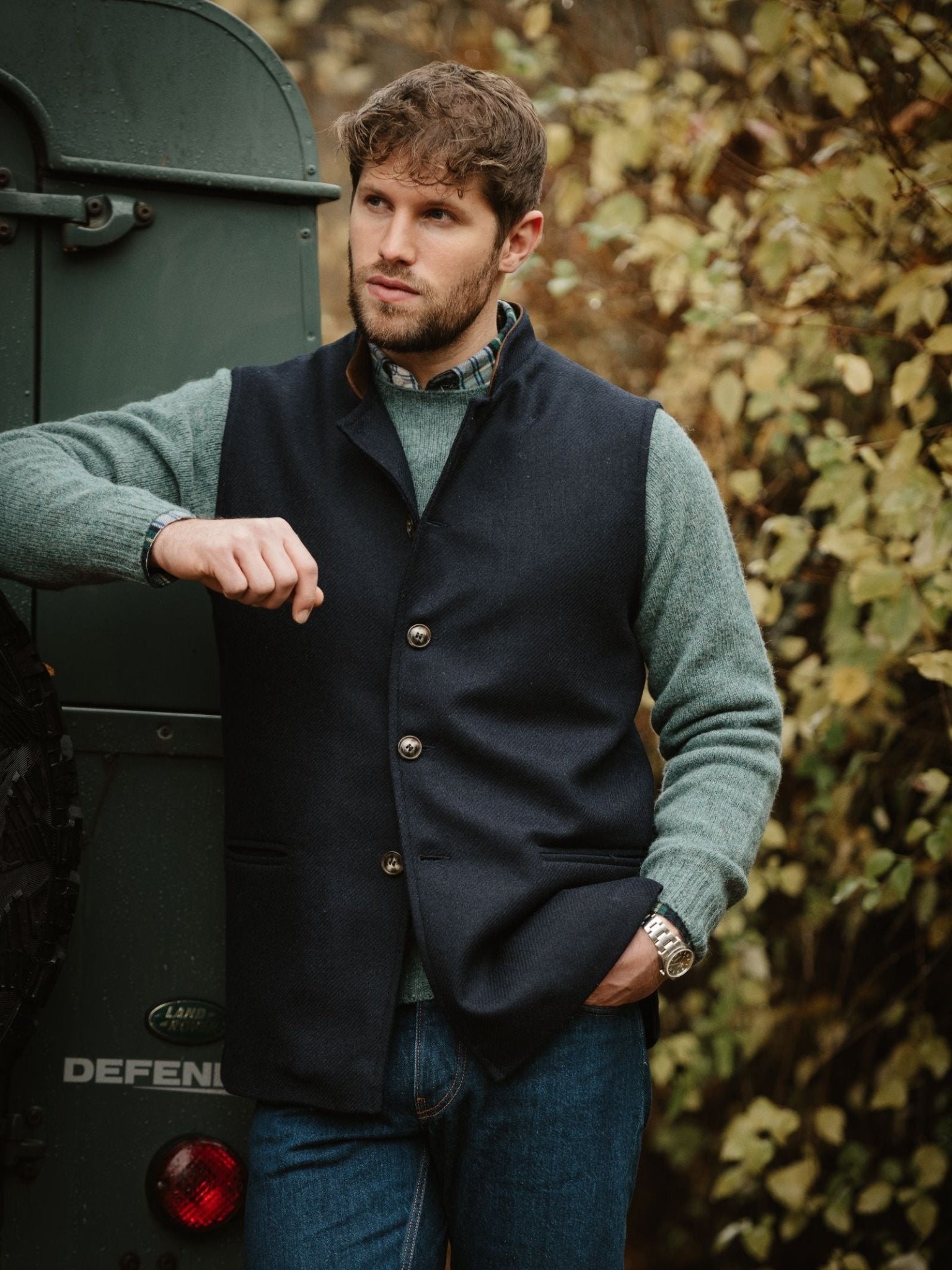 A man with short brown hair and a beard leans against a green Land Rover Defender, wearing the Campbell's of Beauly Tweed Nehru Gilet, green sweater, navy vest, and blue jeans, with autumn foliage in the background.