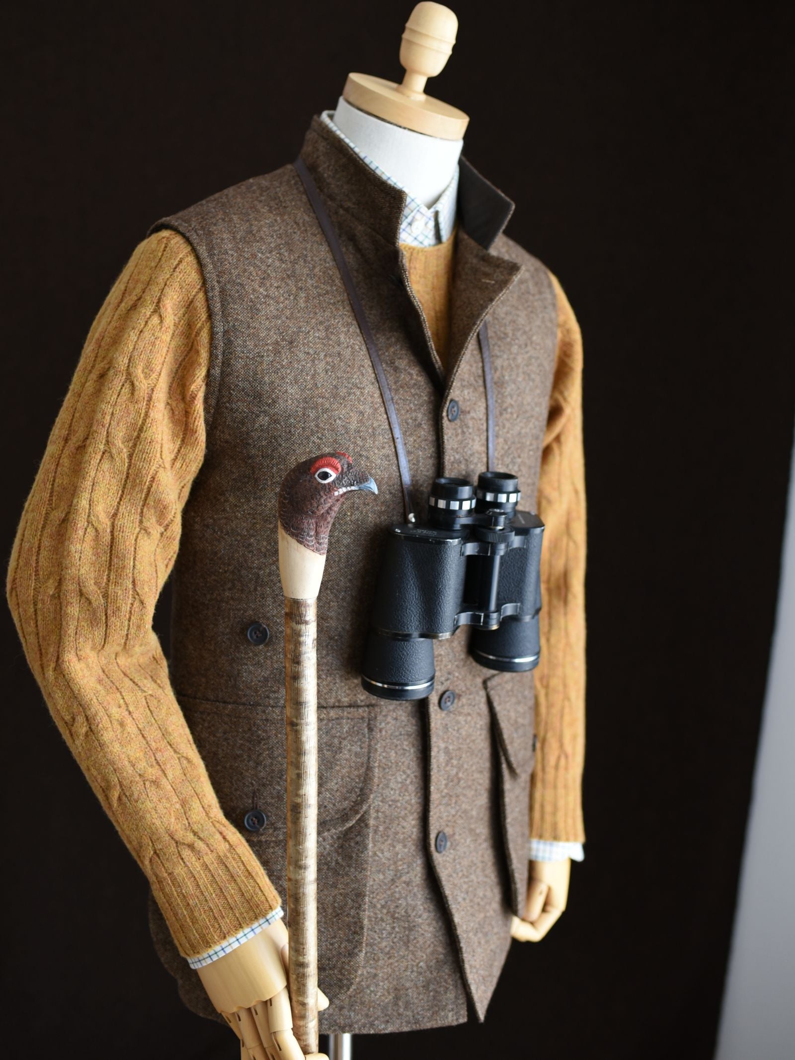A mannequin wears Campbell's of Beauly Auld Stock Field Nehru Gilet over a mustard cable-knit sweater, with binoculars around the neck and a walking stick topped with a bird-shaped handle.