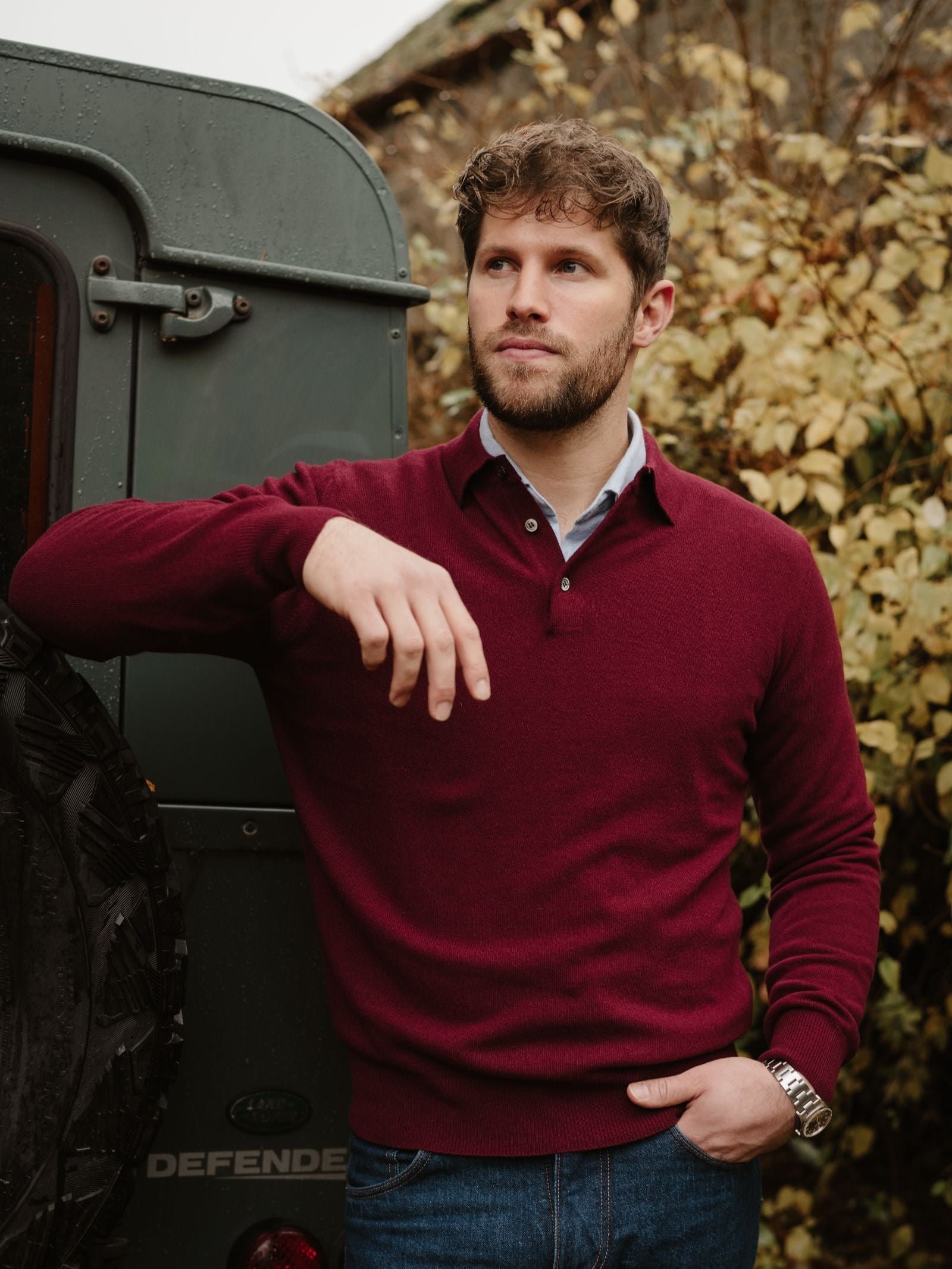 A man with curly hair and a beard, wearing Campbell’s of Beauly’s Merino & Cashmere Rugby sweater and jeans, leans casually against a green Land Rover Defender with autumn foliage in the background.