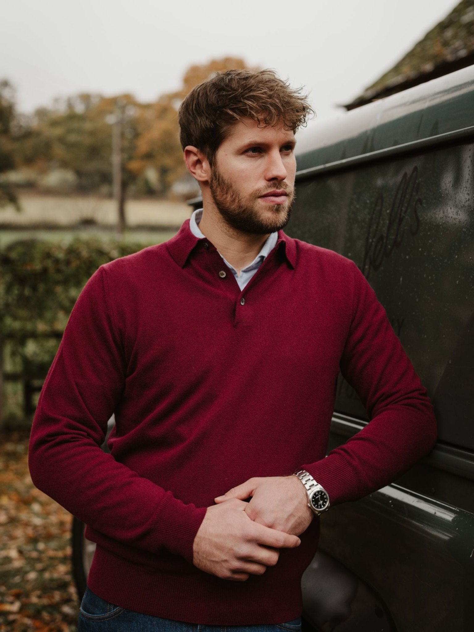 A man with short brown hair and a beard, wearing a maroon Campbell's of Beauly Merino & Cashmere Rugby sweater and a wristwatch, stands outdoors against a dark green vehicle with trees and grass in the background.