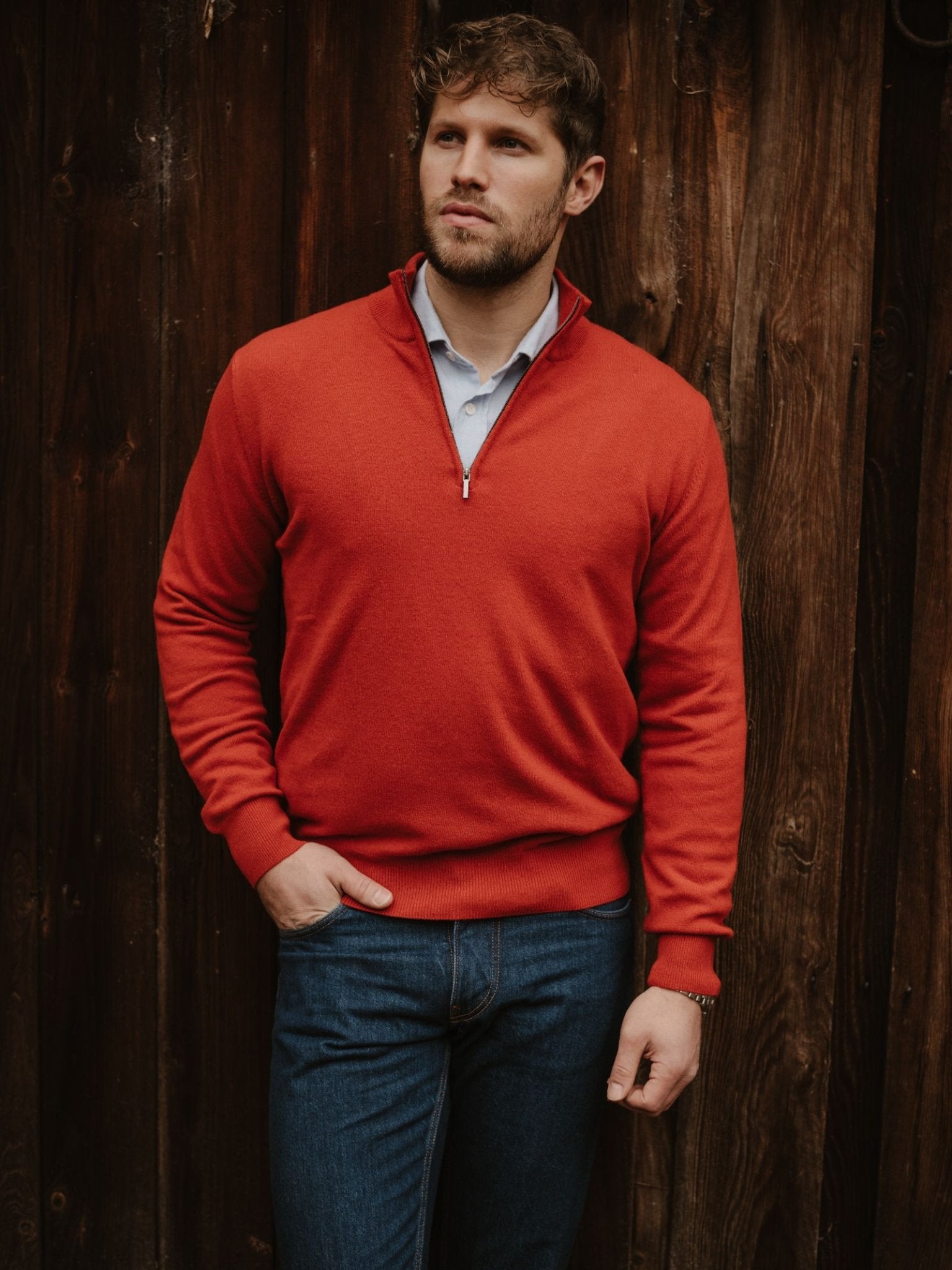 A man with short brown hair and a beard stands by a wooden wall, wearing Campbell's of Beauly Merino & Cashmere Quarter Zip Jumper in bright red over a light blue collared shirt and dark blue jeans, one hand in his pocket.