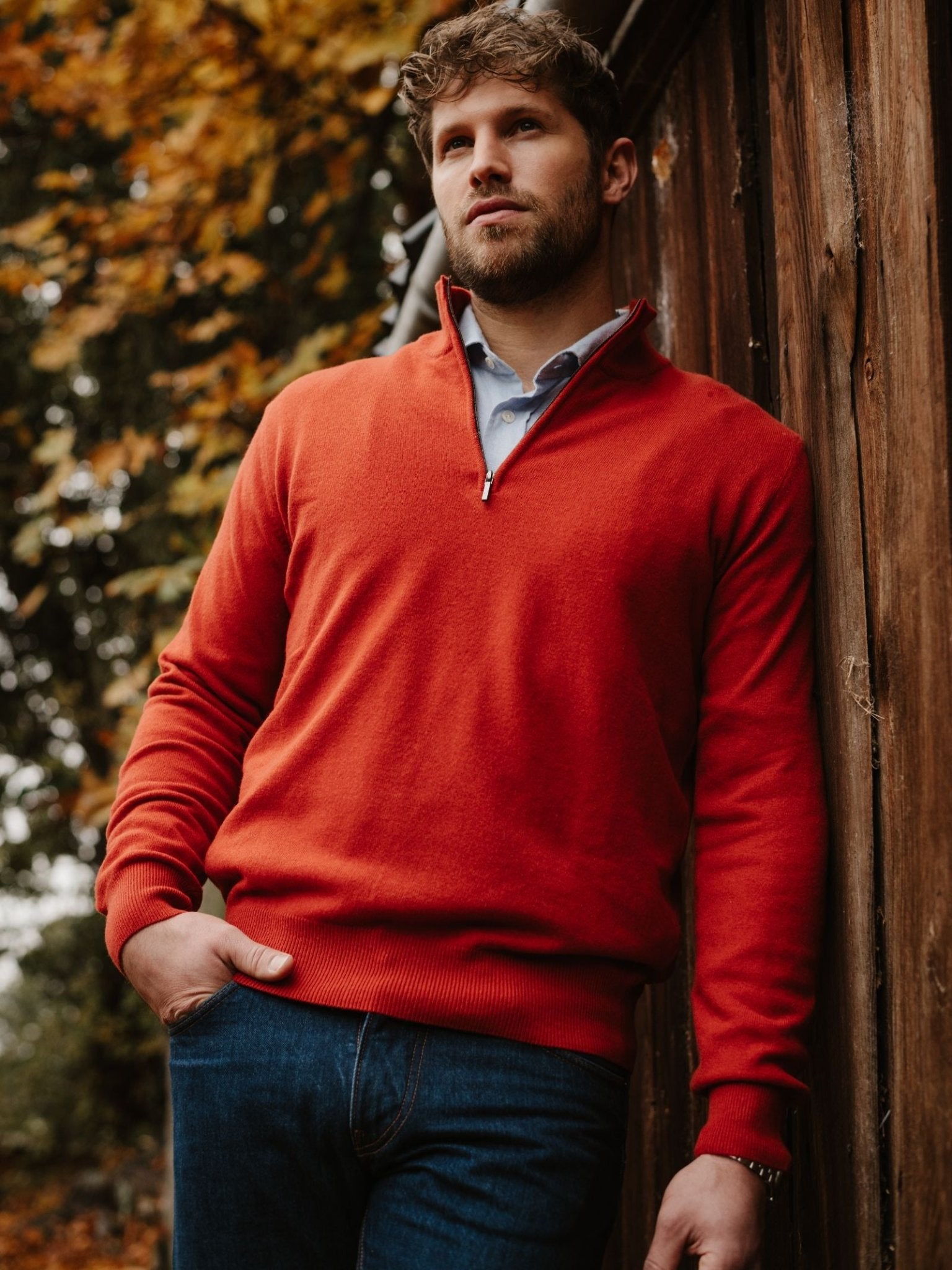A man with curly hair and a beard wears a bright orange Campbell's of Beauly Merino & Cashmere Quarter Zip Jumper over a collared shirt and blue jeans, standing outdoors by a wooden wall with autumn leaves behind him.