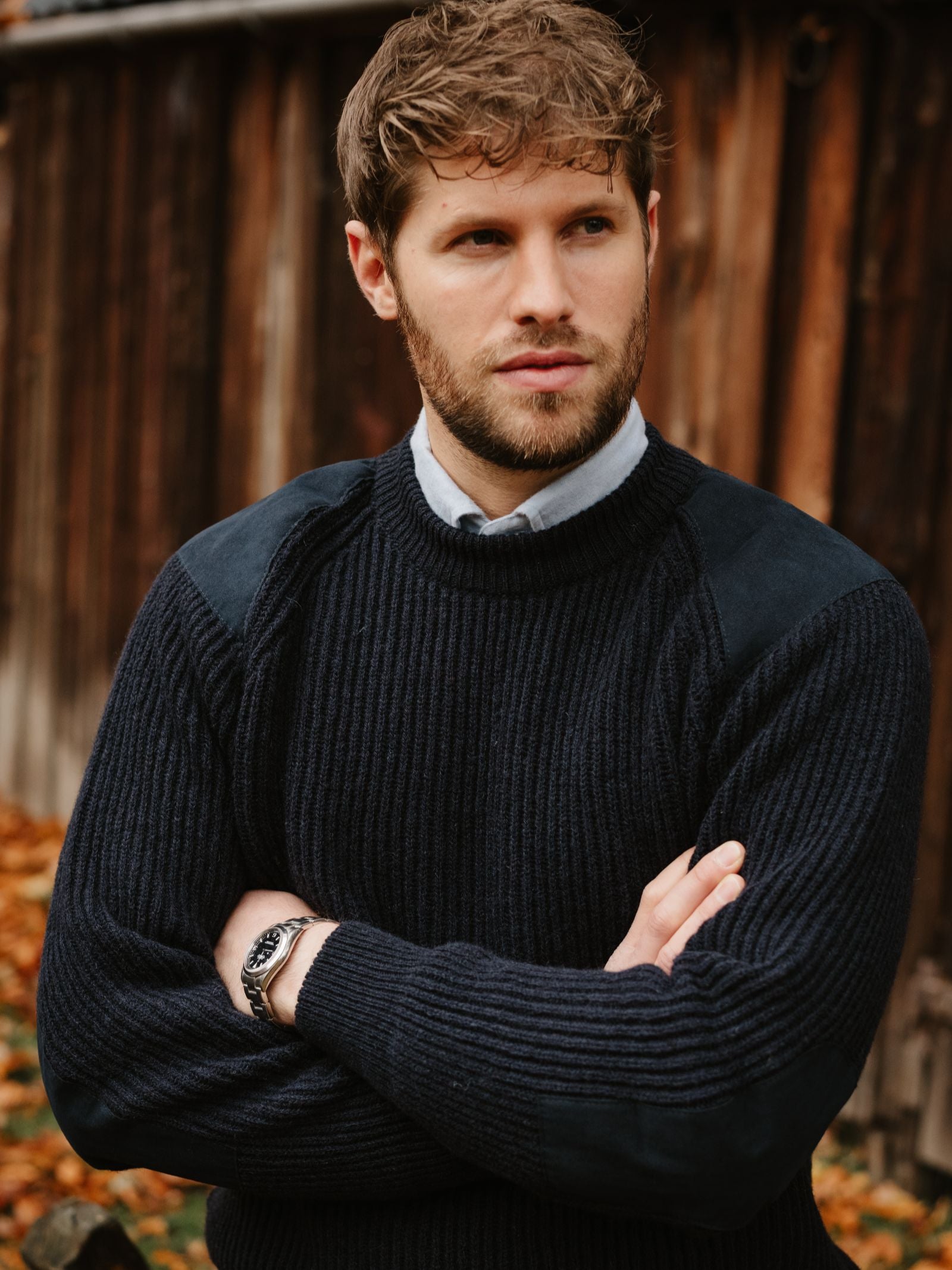A man with light brown hair and a beard stands outside, wearing the Campbell's of Beauly Scotia Suede Jumper over a collared shirt. A rustic wooden wall and autumn leaves complete this timeless outdoor look.