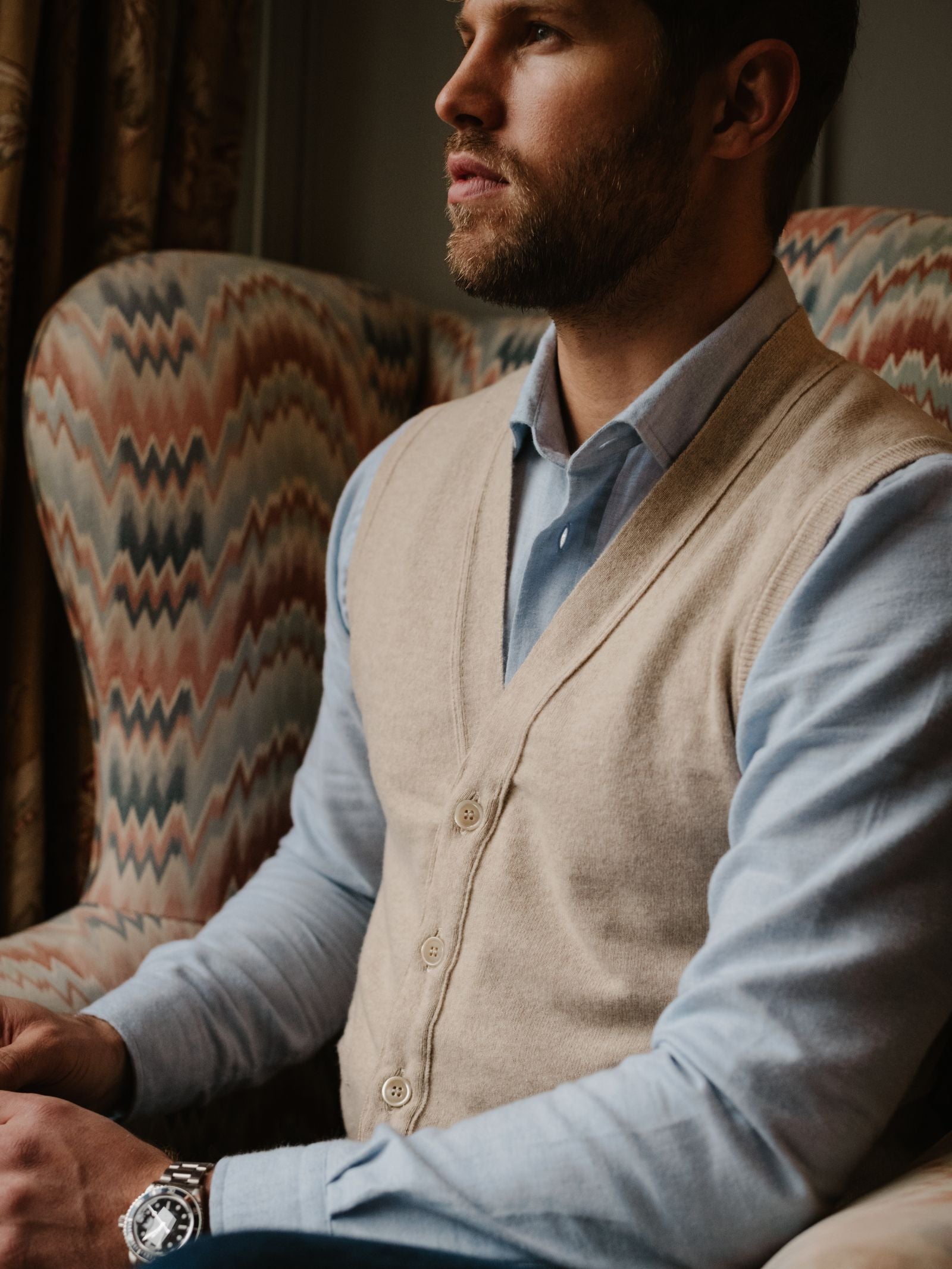A man with a beard, wearing a light blue shirt and the Campbell's of Beauly Geelong Button Waistcoat in beige, sits on a patterned armchair, looking thoughtfully to the side. He also wears a silver watch.