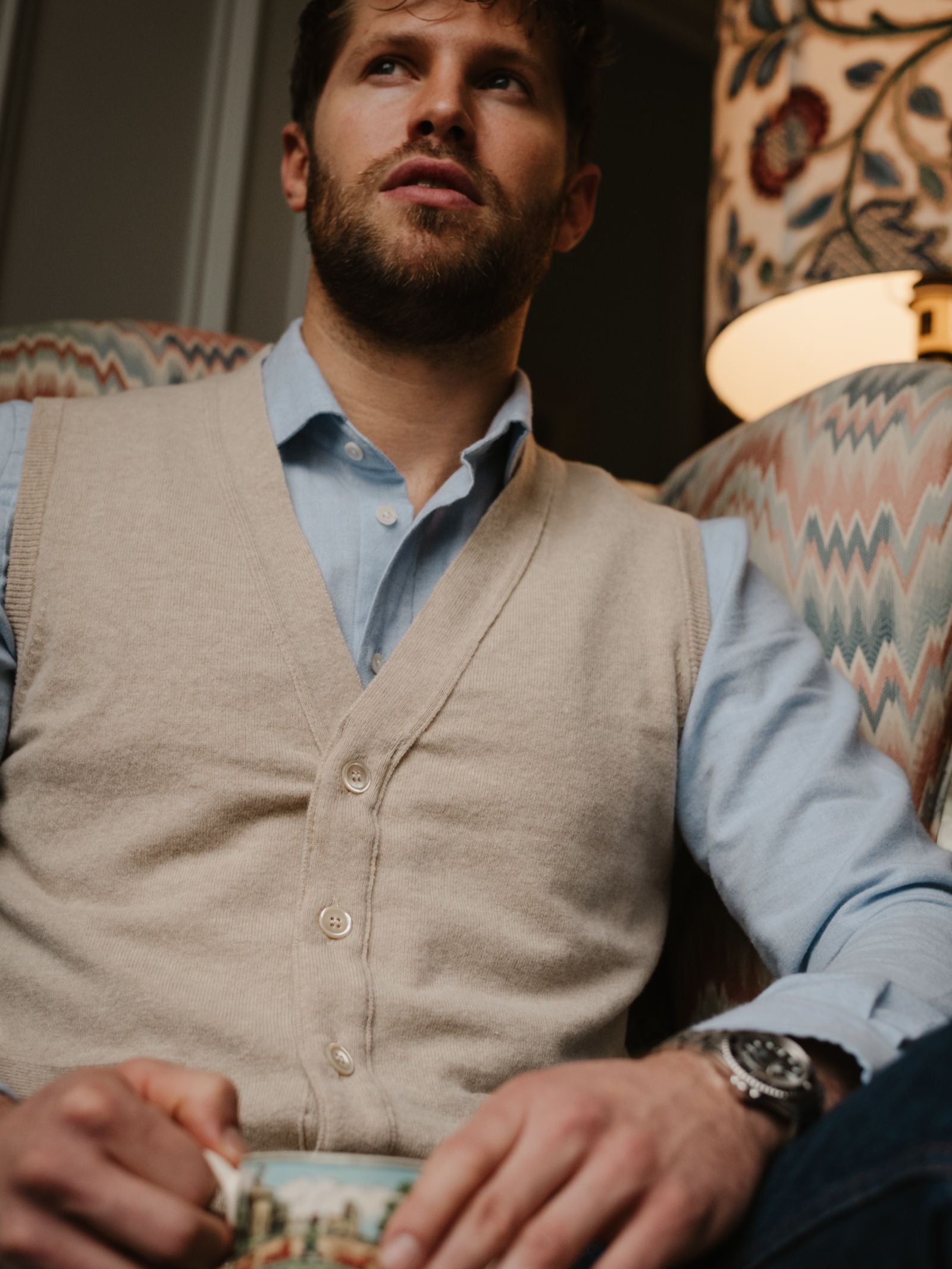 A bearded man in a light blue shirt and Campbell's of Beauly Geelong Button Waistcoat sits on a patterned armchair, holding a book and gazing upward. A patterned lampshade appears in the background.