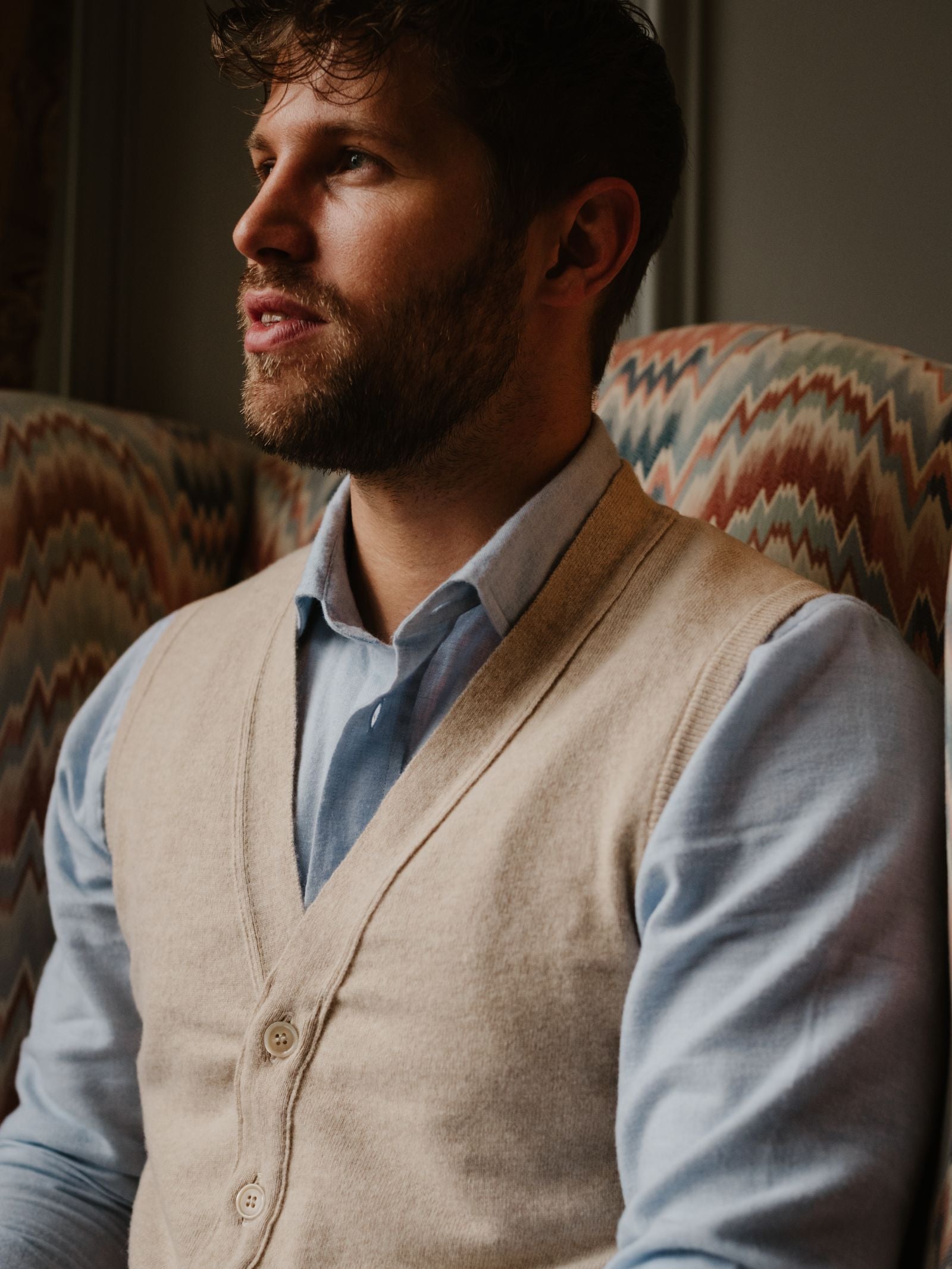 A bearded man with short hair sits on a patterned chair, wearing a light blue shirt and a beige Geelong Button Waistcoat by Campbell's of Beauly, gazing thoughtfully to the side in warm, cozy lighting.