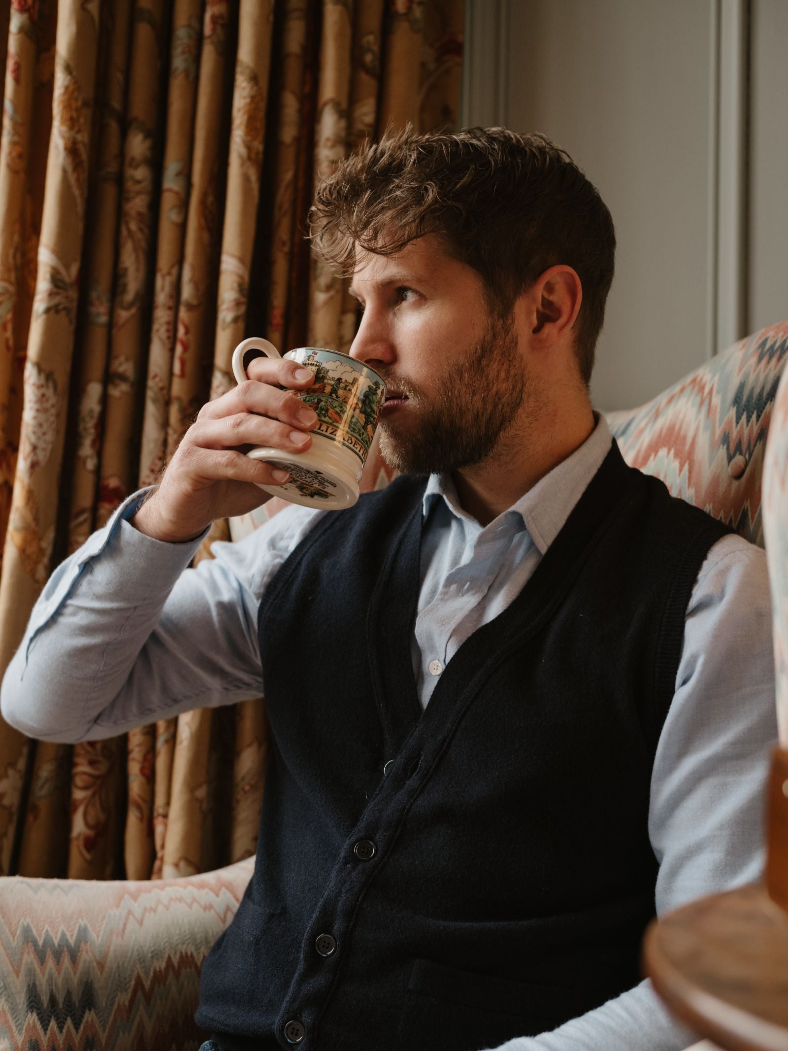 A man with short brown hair and a beard, wearing a light blue shirt and the Geelong Button Waistcoat by Campbell's of Beauly, sits in an armchair sipping from a patterned mug. Floral curtains and a patterned chair are visible in the background.