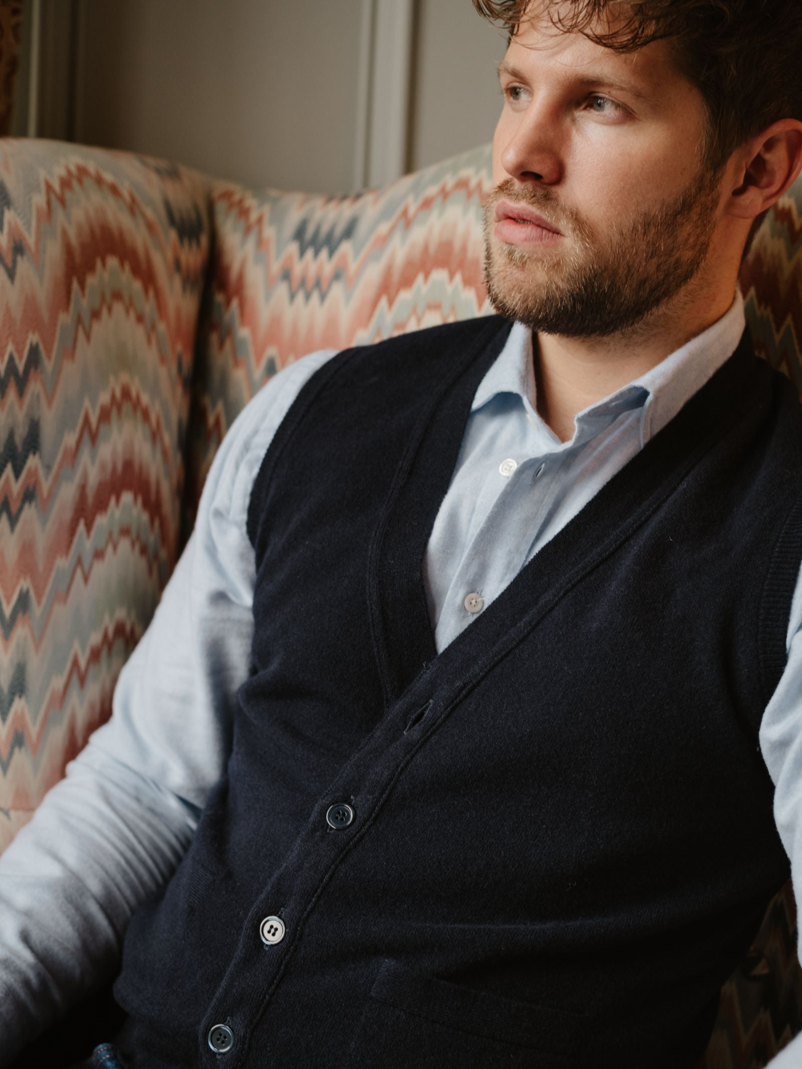 A man with curly hair and a short beard sits on a patterned armchair, wearing a light blue shirt and the Geelong Button Waistcoat by Campbell's of Beauly, looking thoughtfully to the side.