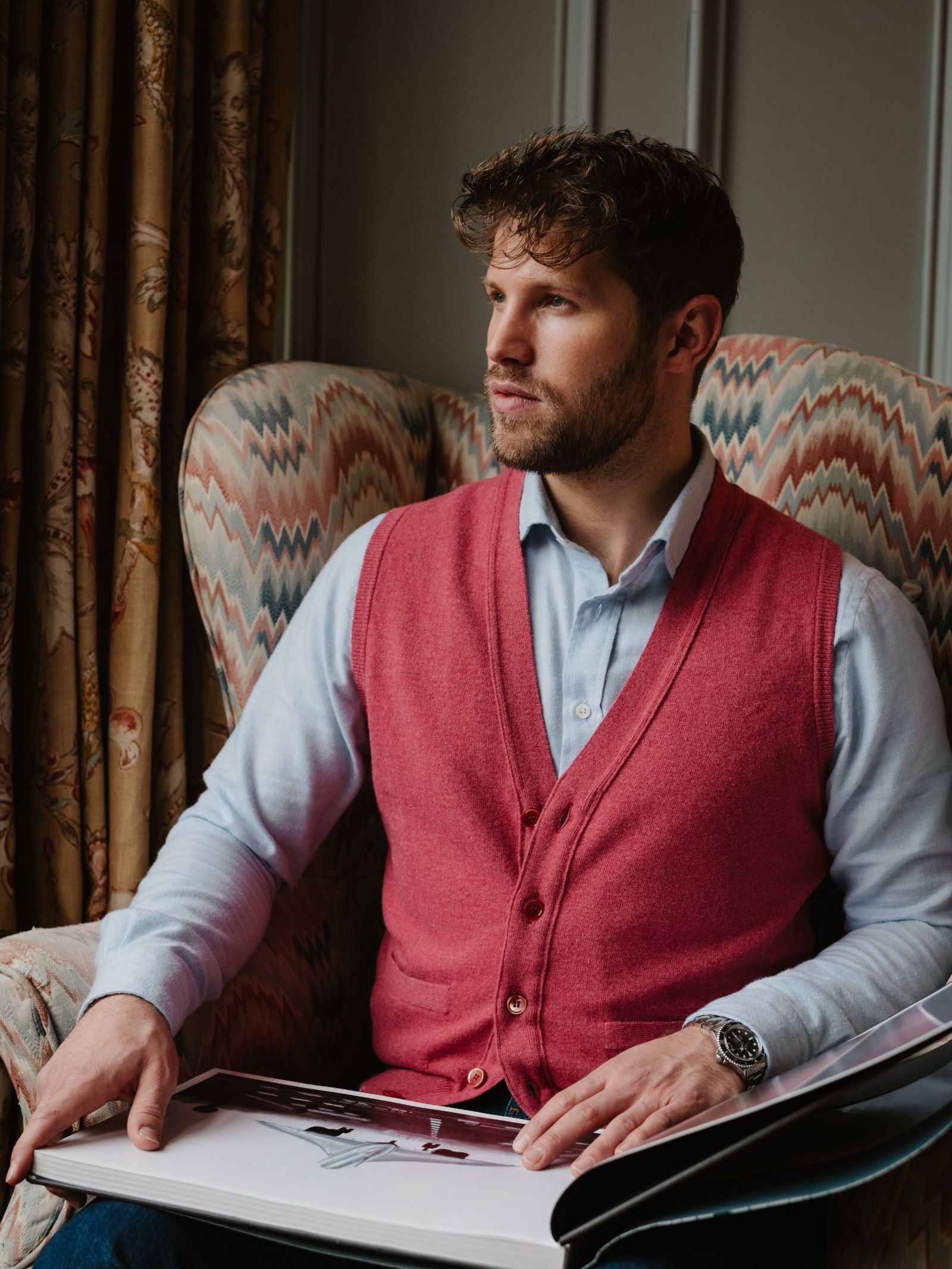 A man with curly hair and a beard sits in an armchair, thoughtfully holding open a large book while wearing a light blue shirt and the Geelong Button Waistcoat by Campbell's of Beauly.