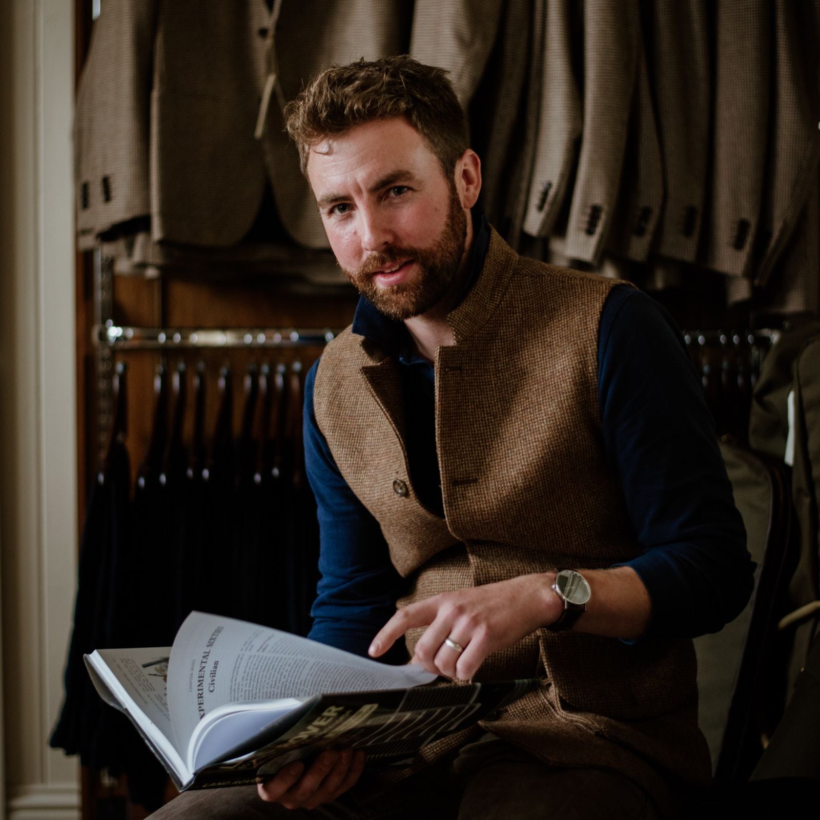 A man with a beard, wearing a brown vest over a dark shirt, sits and looks at the camera while holding and pointing to a page in an open book. Behind him are hanging jackets and suits.