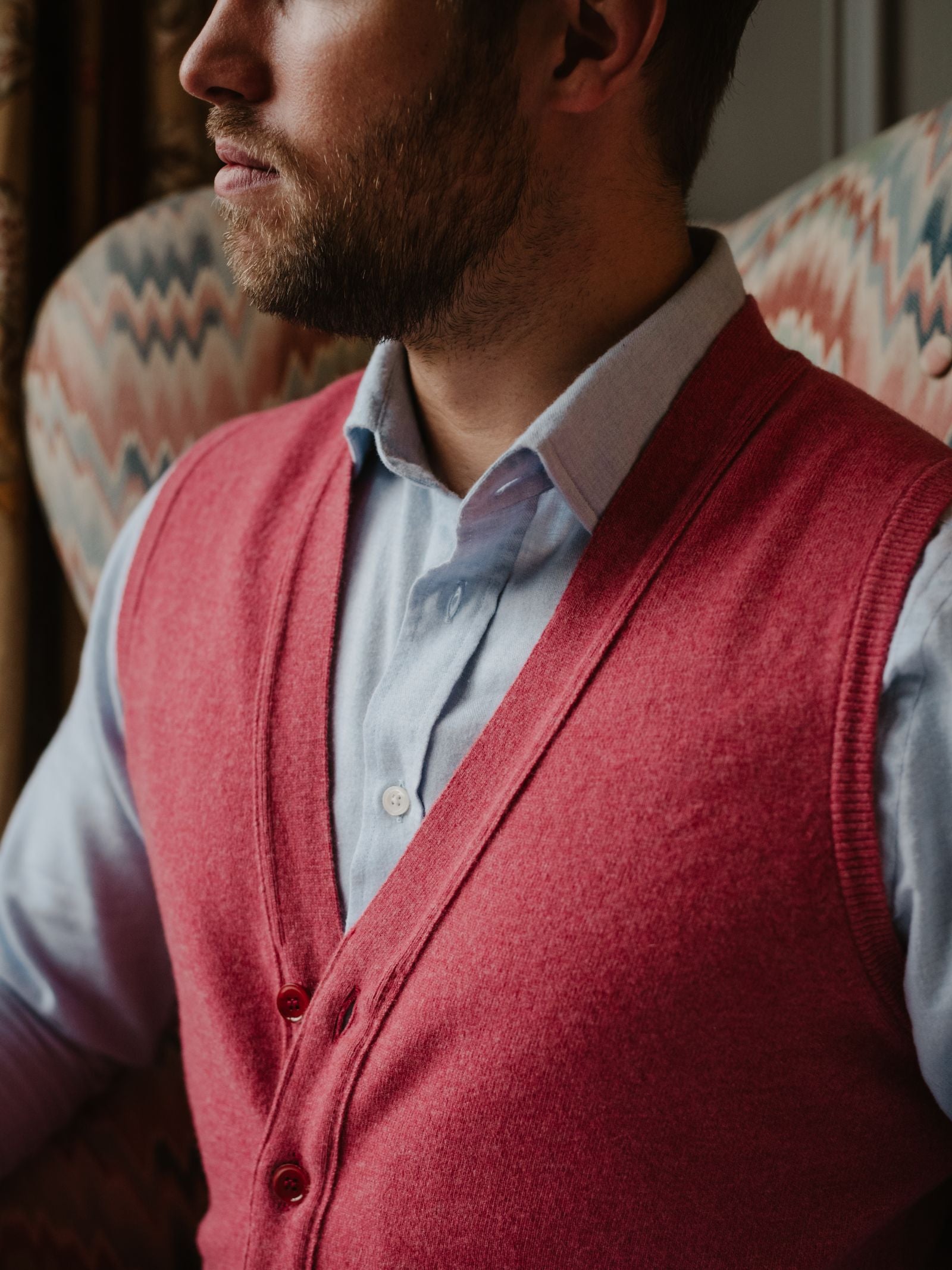 A bearded man wearing a light blue button-up shirt and a red Campbell's of Beauly Geelong Button Waistcoat sits indoors on patterned upholstery, with his face partially out of frame.