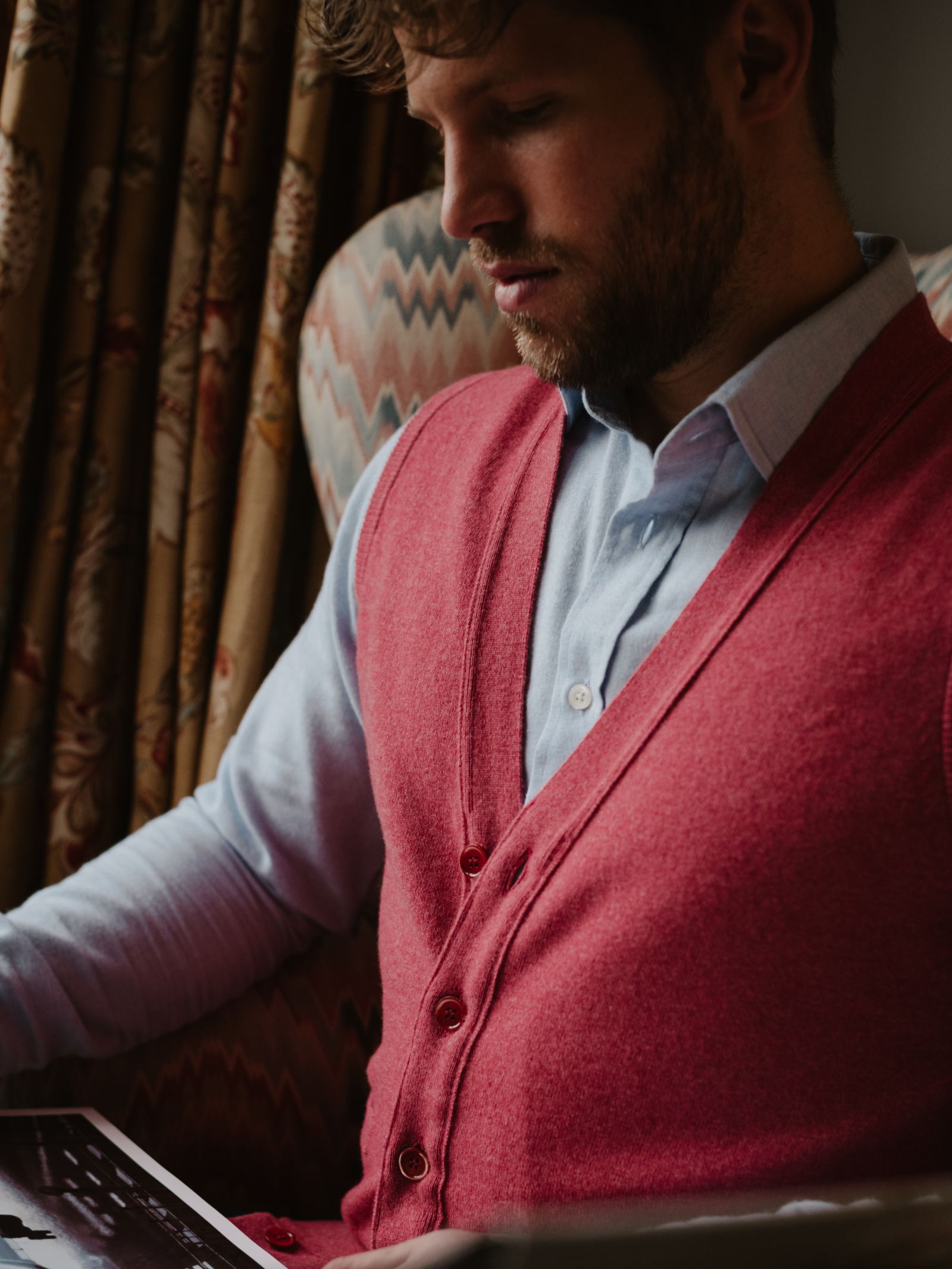 A bearded man sits in a patterned armchair, wearing a light blue dress shirt and the Geelong Button Waistcoat by Campbell's of Beauly. He looks down at an open book, with patterned curtains in the background.