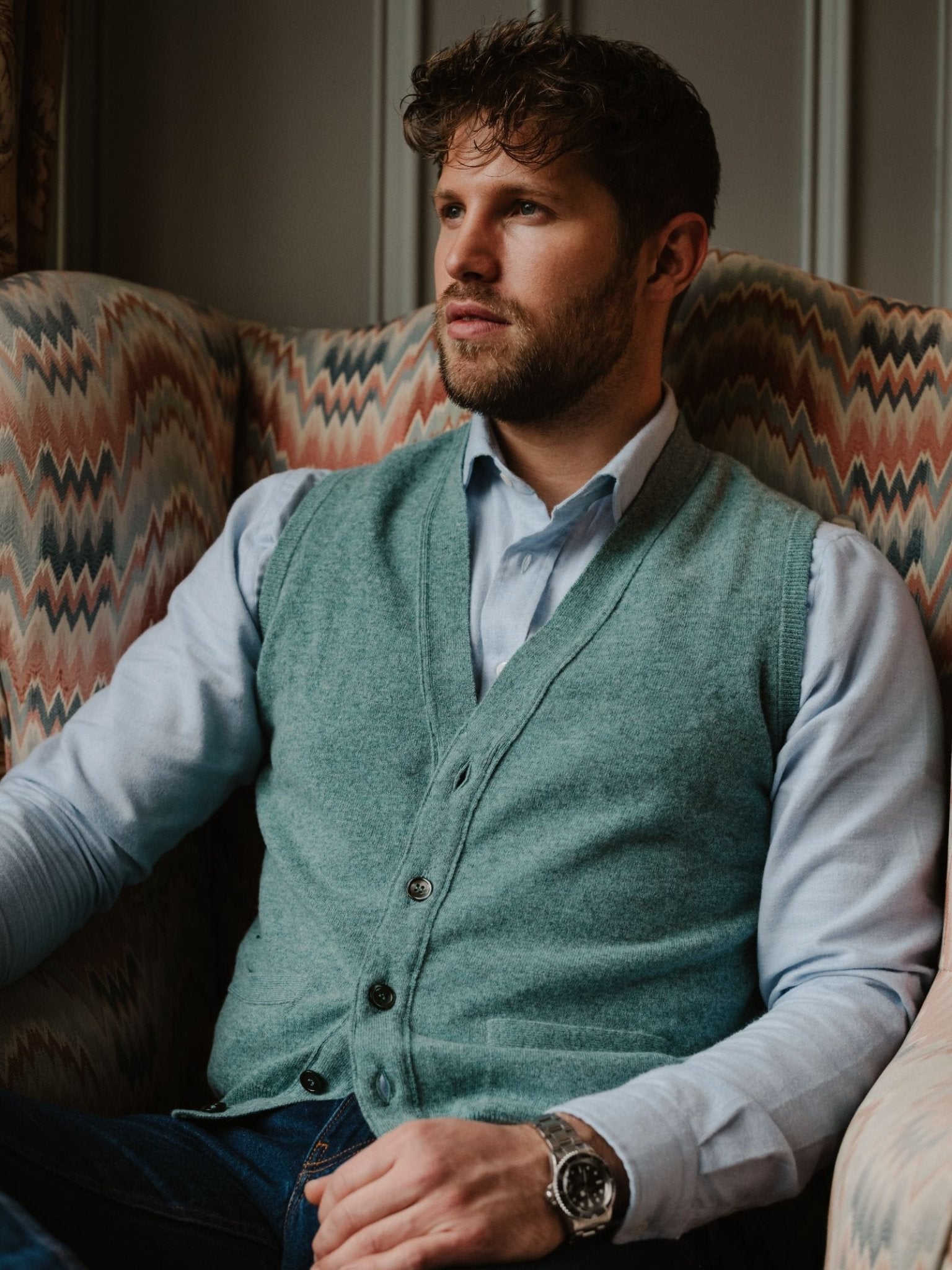 A man with wavy hair and a beard sits in a patterned armchair, wearing a light blue shirt and the Geelong Button Waistcoat by Campbell's of Beauly, gazing thoughtfully into the distance.