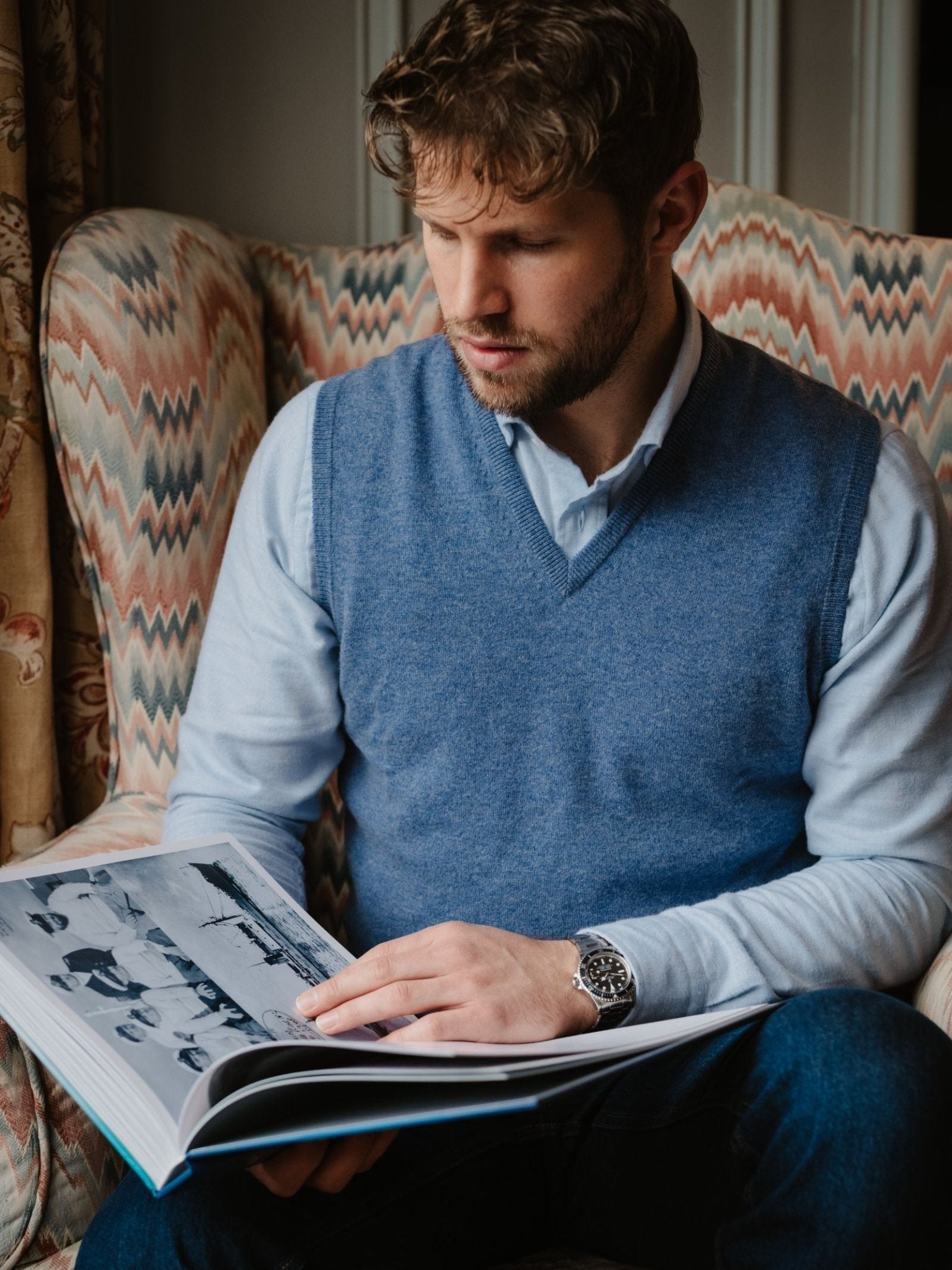 A man with short brown hair and a beard sits in an armchair, wearing the Campbell's of Beauly Auld Stock Cashmere Slipover over a light blue shirt, looking at a large open photo book with black-and-white images.
