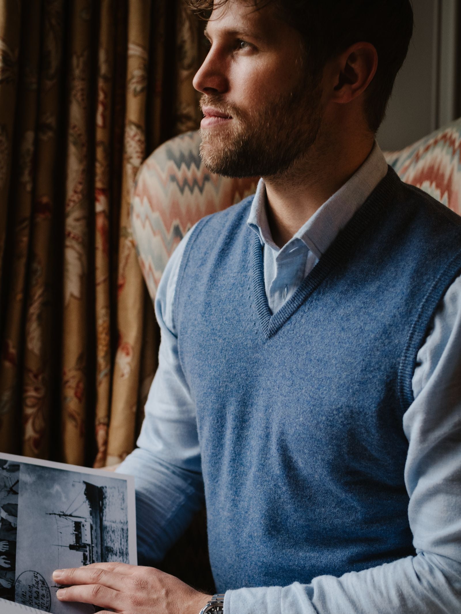 A bearded man in a light blue shirt and an Auld Stock Cashmere Slipover by Campbell's of Beauly sits in a patterned armchair, holding a black-and-white photo and gazing thoughtfully out the window.