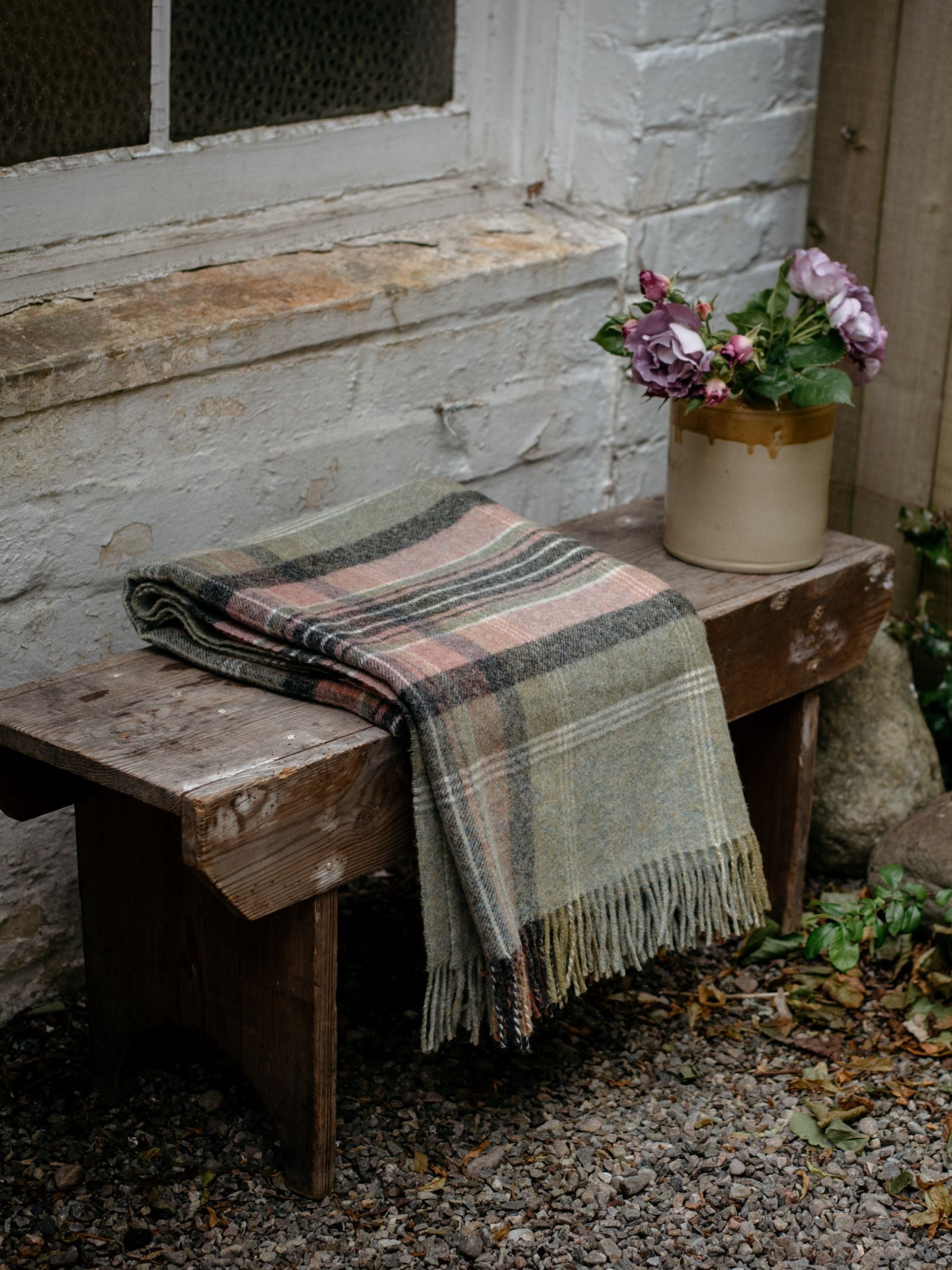 The Inchmore Lambswool Throw by Campbell’s of Beauly, folded with its fringe showing, adds Yorkshire charm on a rustic bench beside a mauve-flowered vase, set against a white brick wall and window.