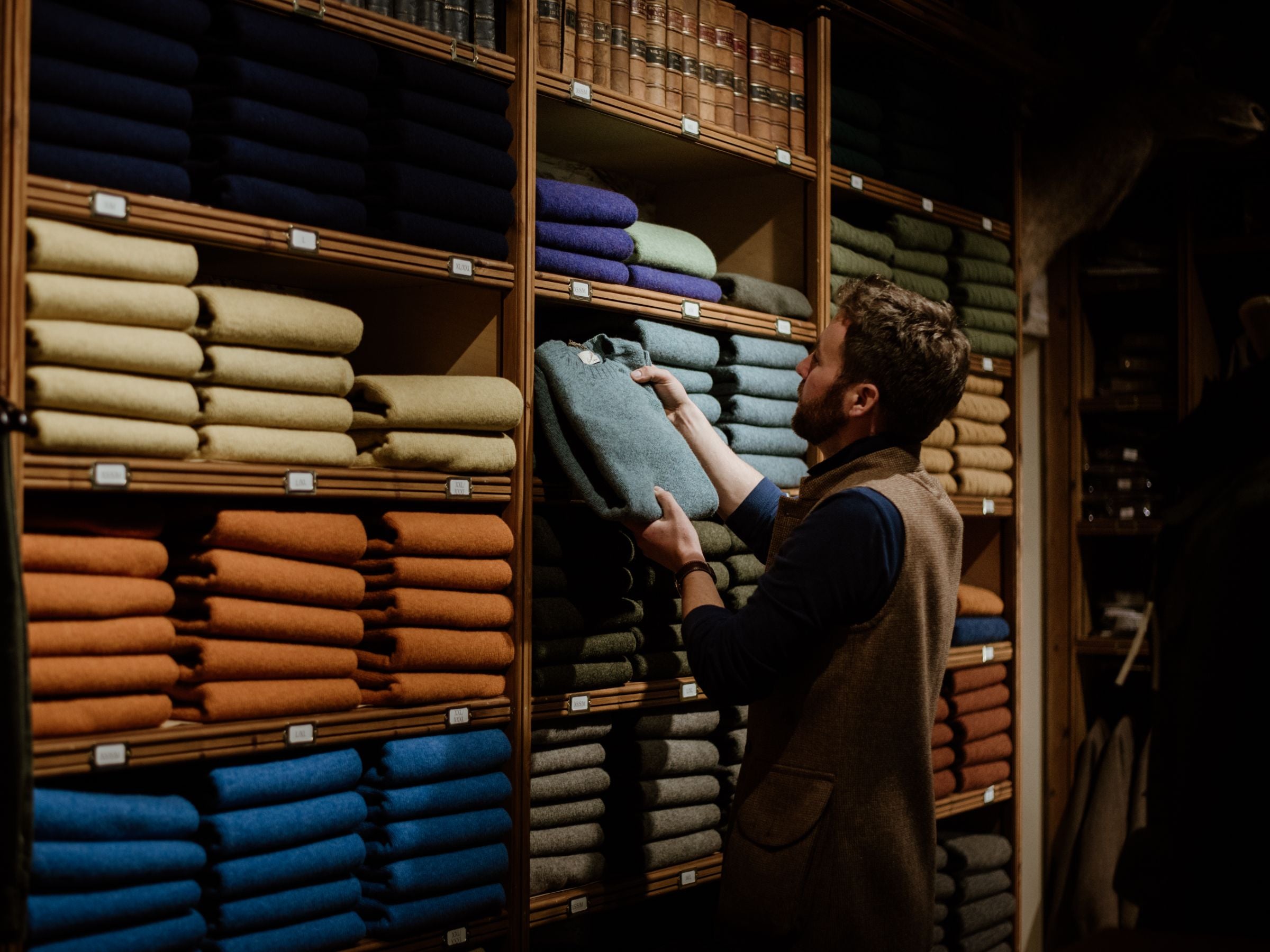 A man stands in front of shelves neatly stacked with colorful sweaters, reaching to take a green sweater from the middle shelf in a cozy, well-organized store.
