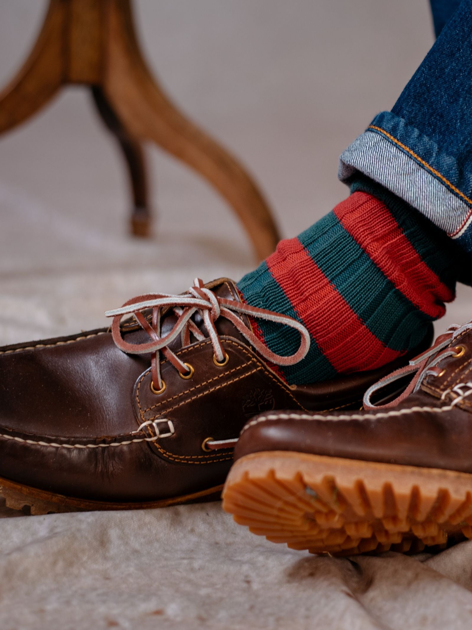 A close-up shows a person wearing Campbell's of Beauly Stripe Socks—green and red cotton socks with reinforced heels—paired with brown leather shoes and blue jeans, with a wooden furniture leg in the background.