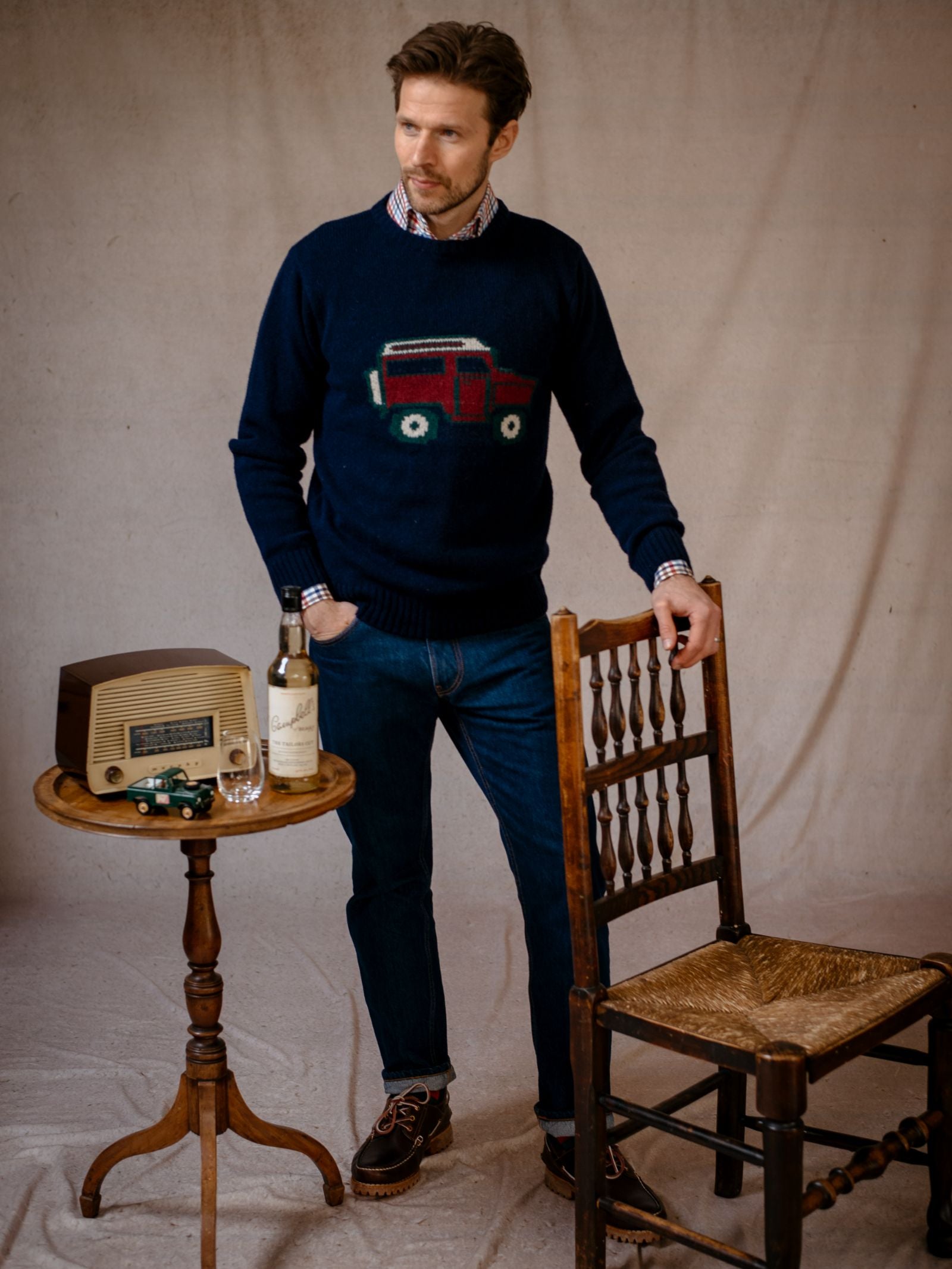 A man in classic knitwear—a navy Unisex Land Rover Defender Jumper by Campbell's of Beauly with a red car design—jeans, and brown shoes stands indoors beside a wooden chair and table holding motoring-themed accessories.