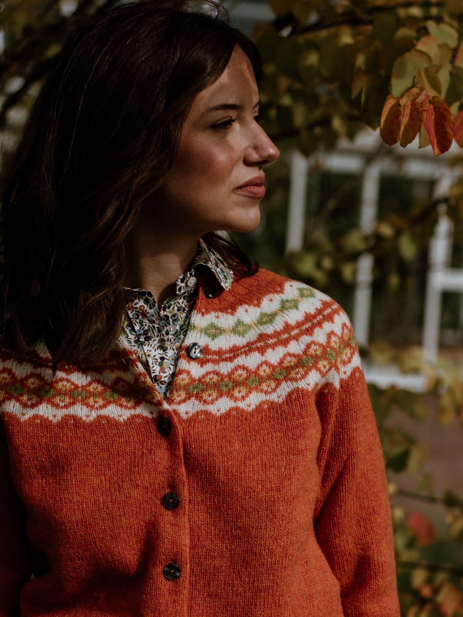 A woman with long brown hair, wearing the Campbell's of Beauly Auld Stock Shetland Fairisle Cardigan over a floral shirt, stands outdoors amid autumn leaves, soft sunlight highlighting her Scottish knitwear.