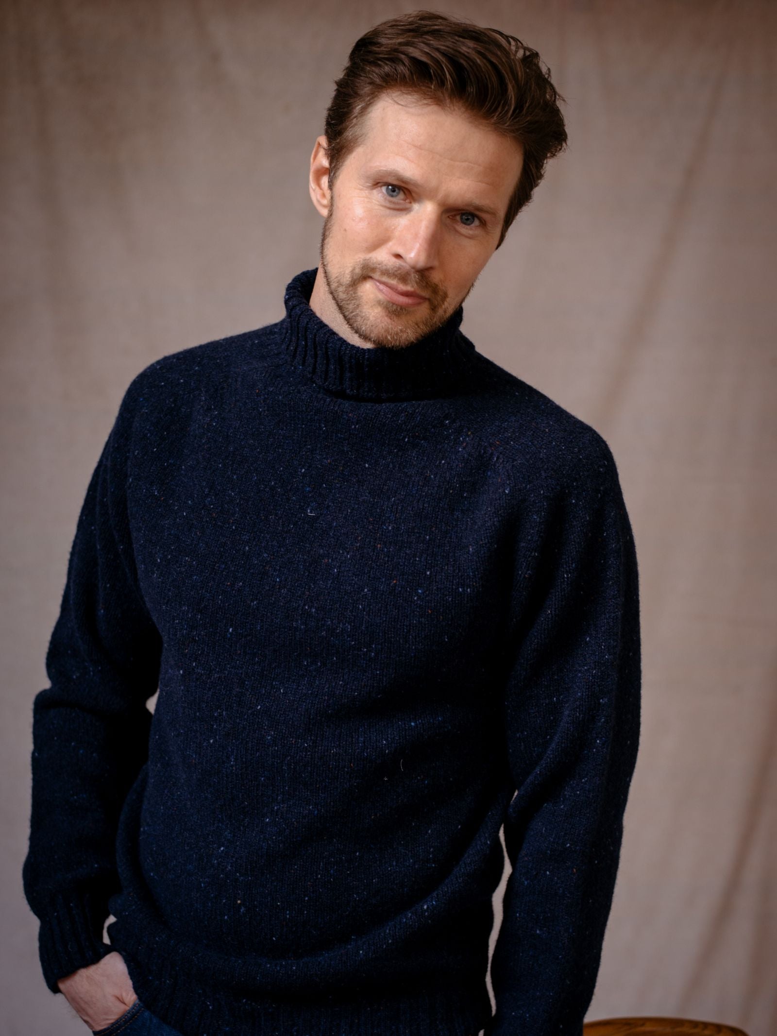 A man with short brown hair and a beard wears the Campbell's of Beauly Donegal Lambswool Roll Neck in dark blue. He stands against a neutral beige background, looking at the camera with a slight smile and one hand in his pocket.