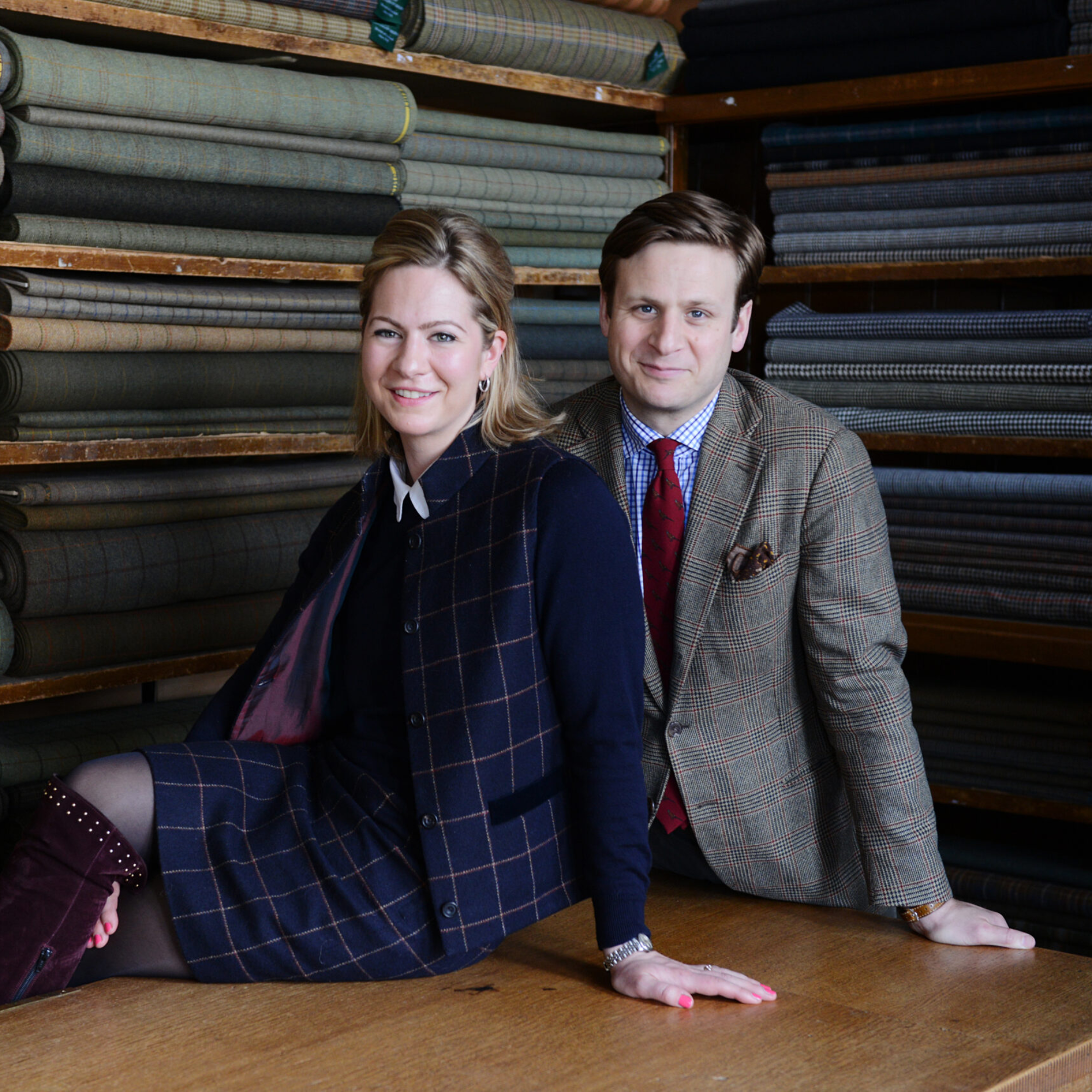 A woman and a man sit on a wooden table in a fabric shop, both dressed in smart, classic plaid outfits. Rolls of fabric are neatly stacked on shelves behind them. They are smiling and looking at the camera.