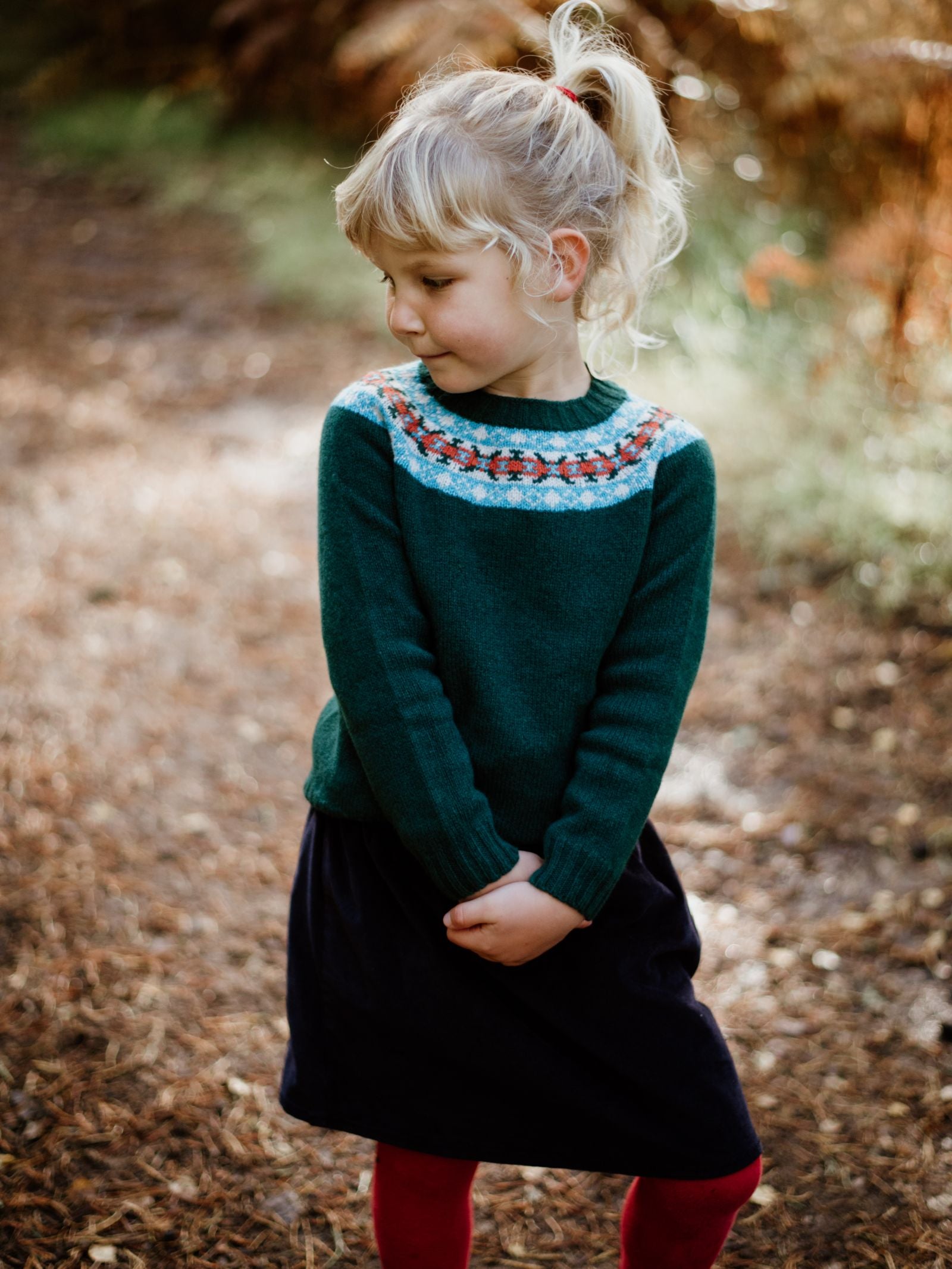 A young blonde girl with her hair in a ponytail looks down and to the left, wearing Campbell's of Beauly's Auld Stock Mini Fairisle Crew Jumper in dark green, expertly knitted in Scotland from soft Geelong lambswool.