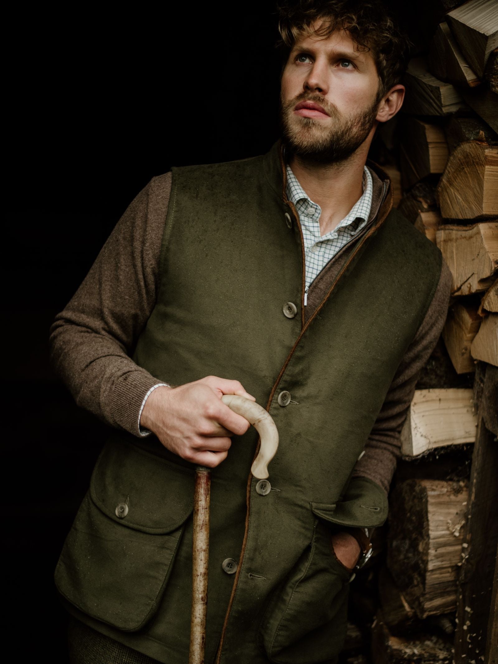 A bearded man with curly hair stands by stacked wood, holding a walking stick and gazing upward, wearing Campbell's of Beauly's Auld Stock Field Nehru Gilet in green over a brown shirt.