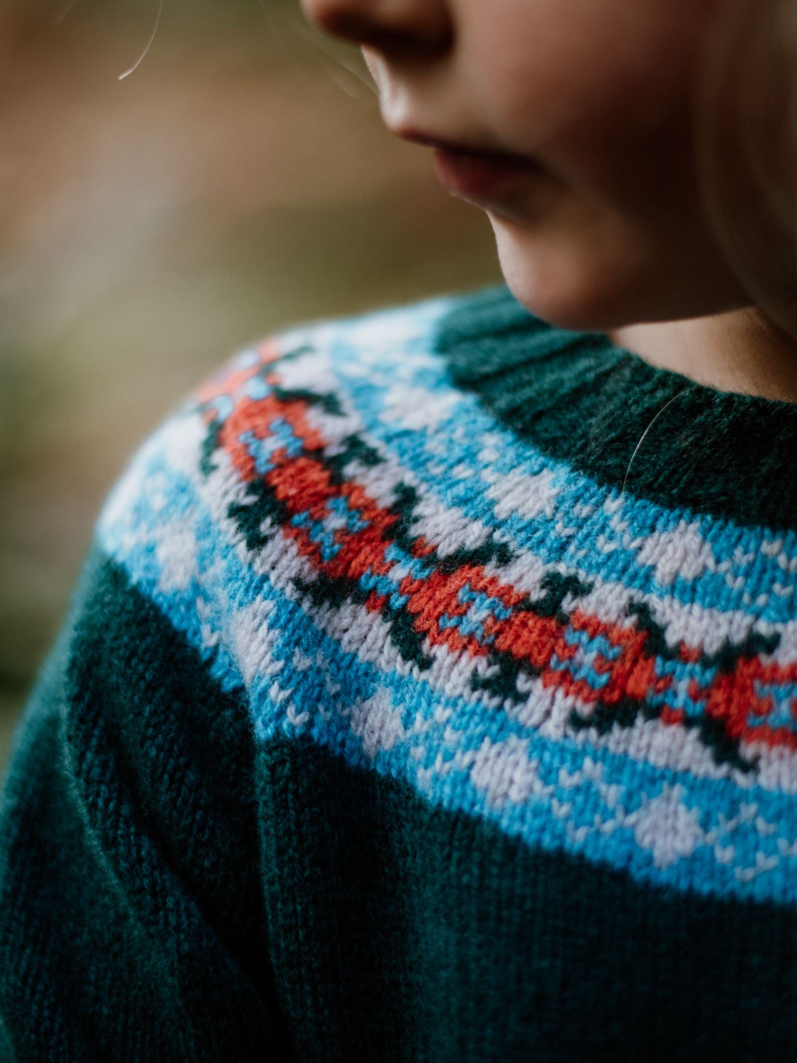 A close-up shows a childs face and shoulder wearing the Campbells of Beauly Mini Fairisle Crew Jumper, featuring a red, blue, and white traditional pattern on green knit lambswool. Only the childs lips and part of their cheek are visible.