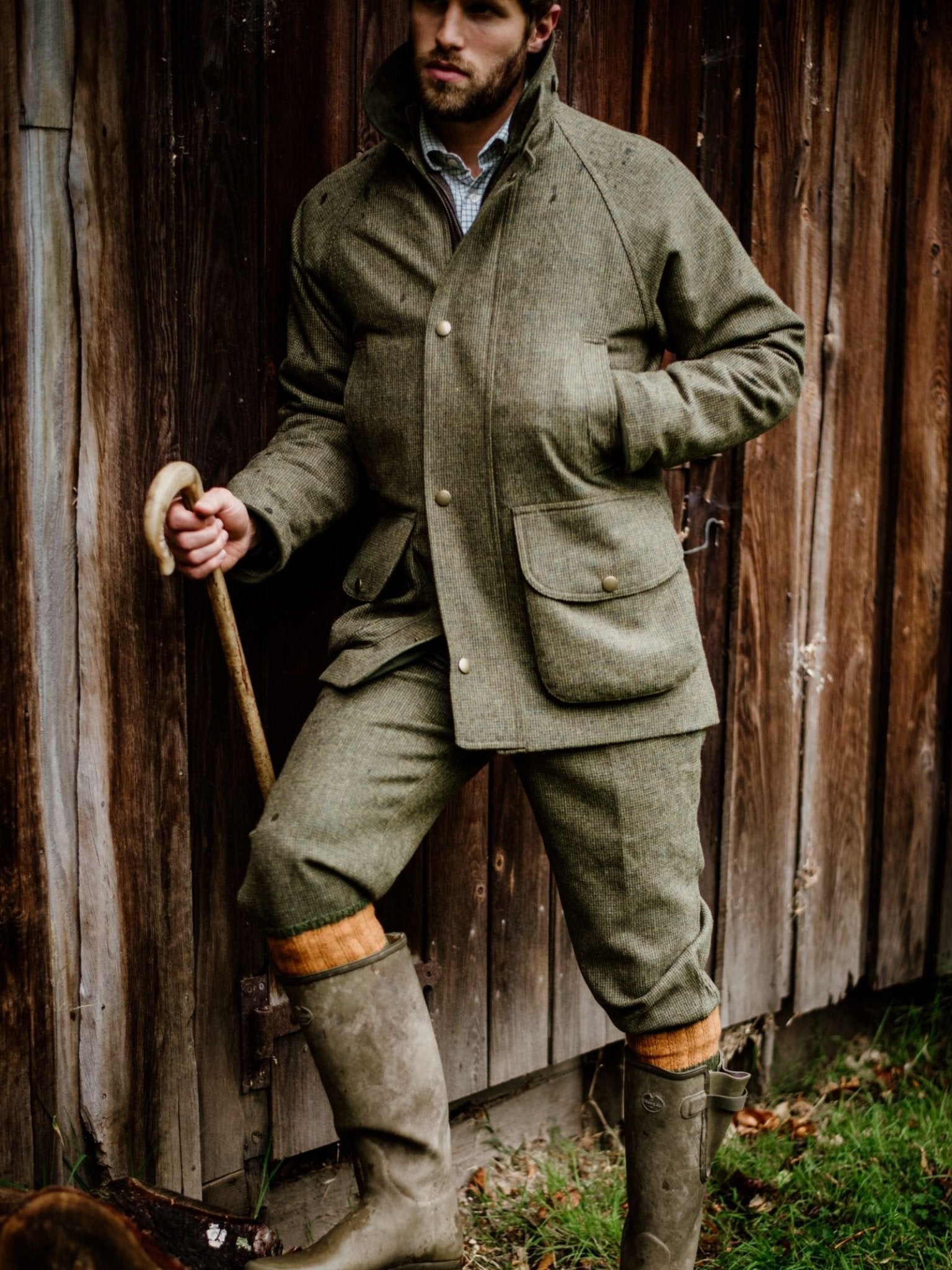 A man in Campbell's of Beauly Tweed Breeks and a green Scottish-woven tweed jacket leans against a rustic wooden wall, holding a cane. He wears tall rubber boots and orange socks, embodying classic countryside style.