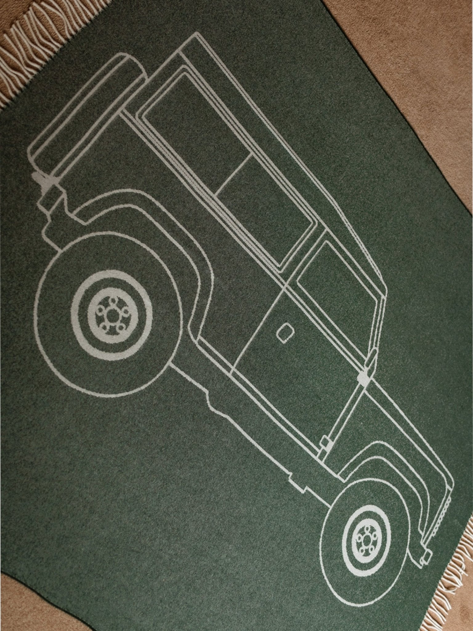 The Land Rover Defender Jacquard Large Scale Motif rug by Campbell's of Beauly, in green with cream fringes, shows a white outline of a four-door off-road vehicle and is displayed on a tan carpeted floor.
