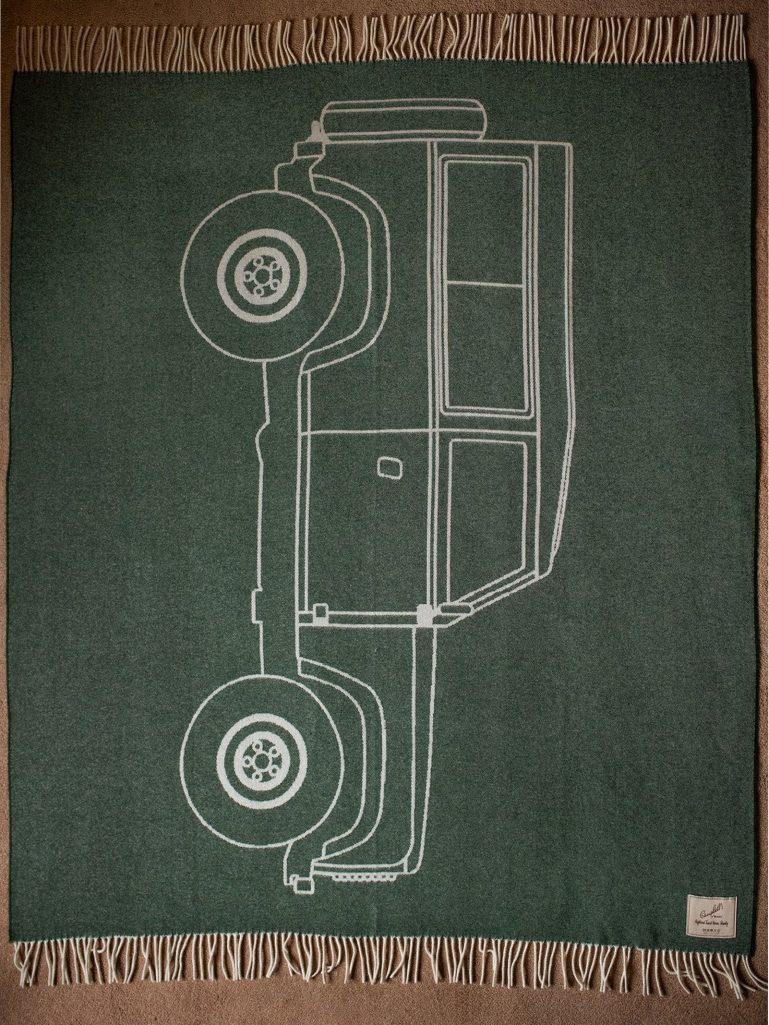 The Campbell's of Beauly Land Rover Defender Jacquard Large Scale Motif is a green wool blanket with beige fringe, featuring a white side-outline of the classic off-road vehicle and a small fabric label in one corner.