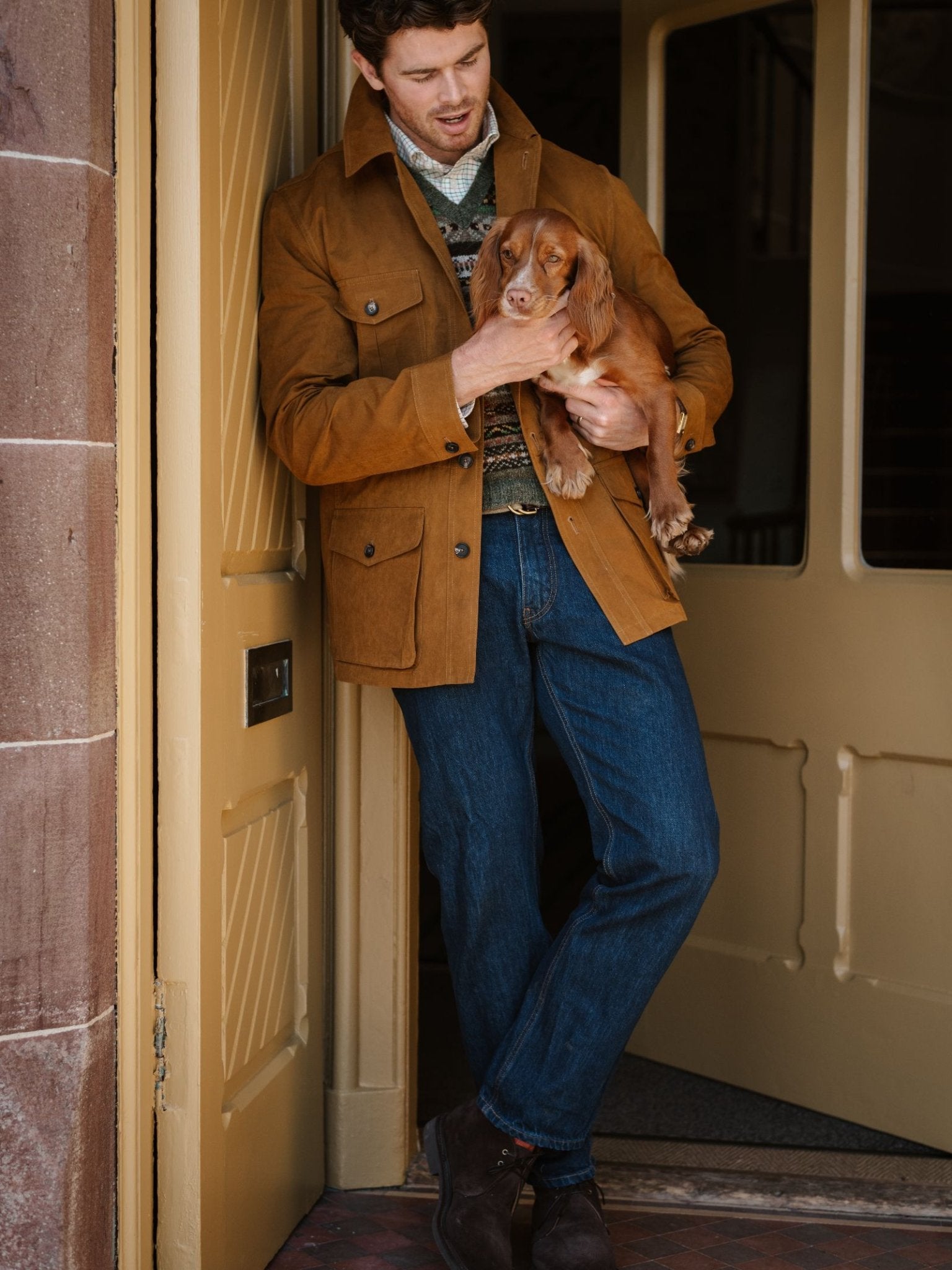 A man wearing Campbell's of Beauly Denim Jeans stands in a doorway, looking down and holding a small brown dog in his arms.