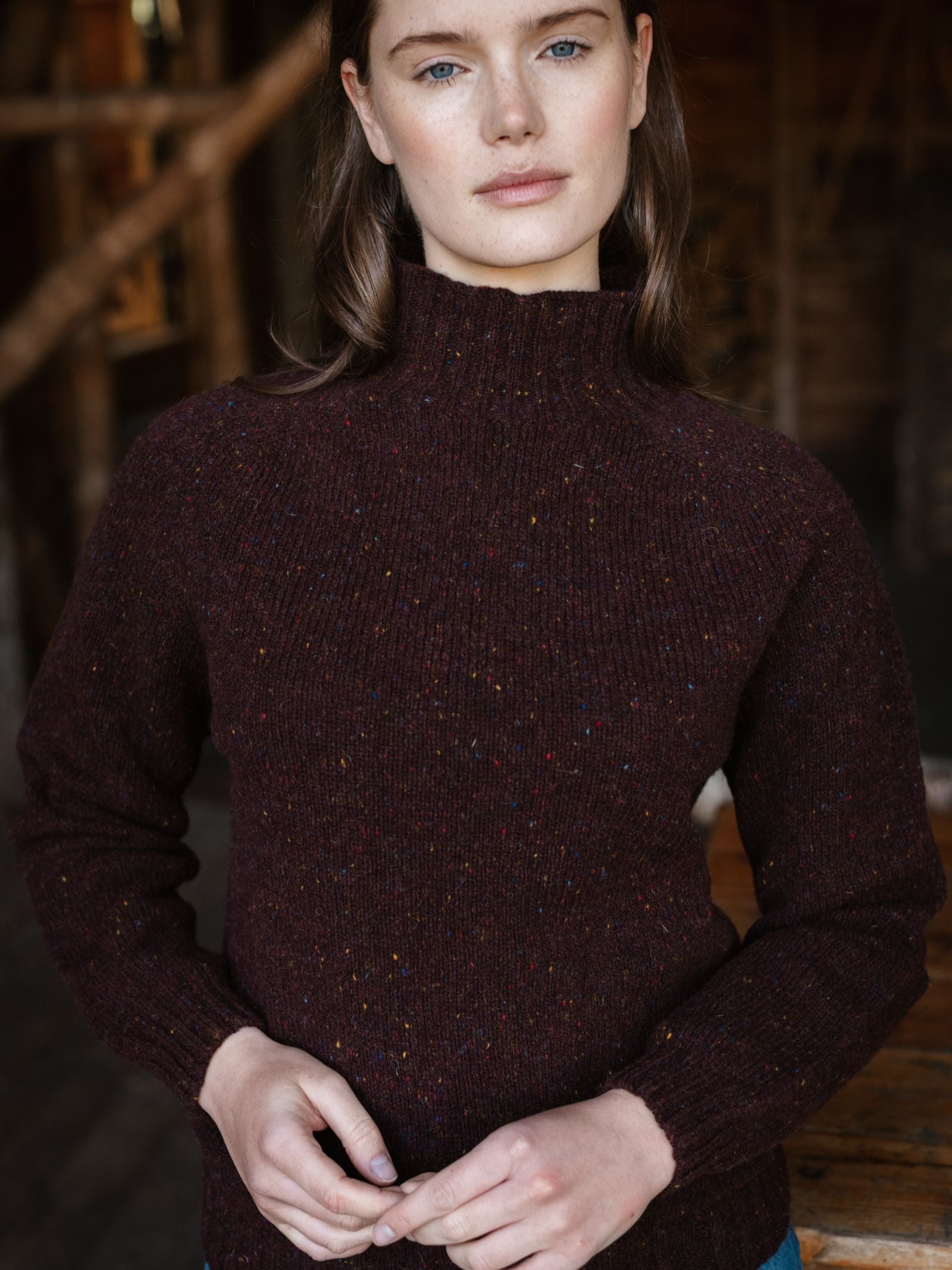 A woman with long brown hair and blue eyes stands indoors, wearing the Campbell's of Beauly Donegal Lambswool Funnel Neck Jumper in dark maroon, her hands gently clasped as she looks at the camera with a neutral expression.